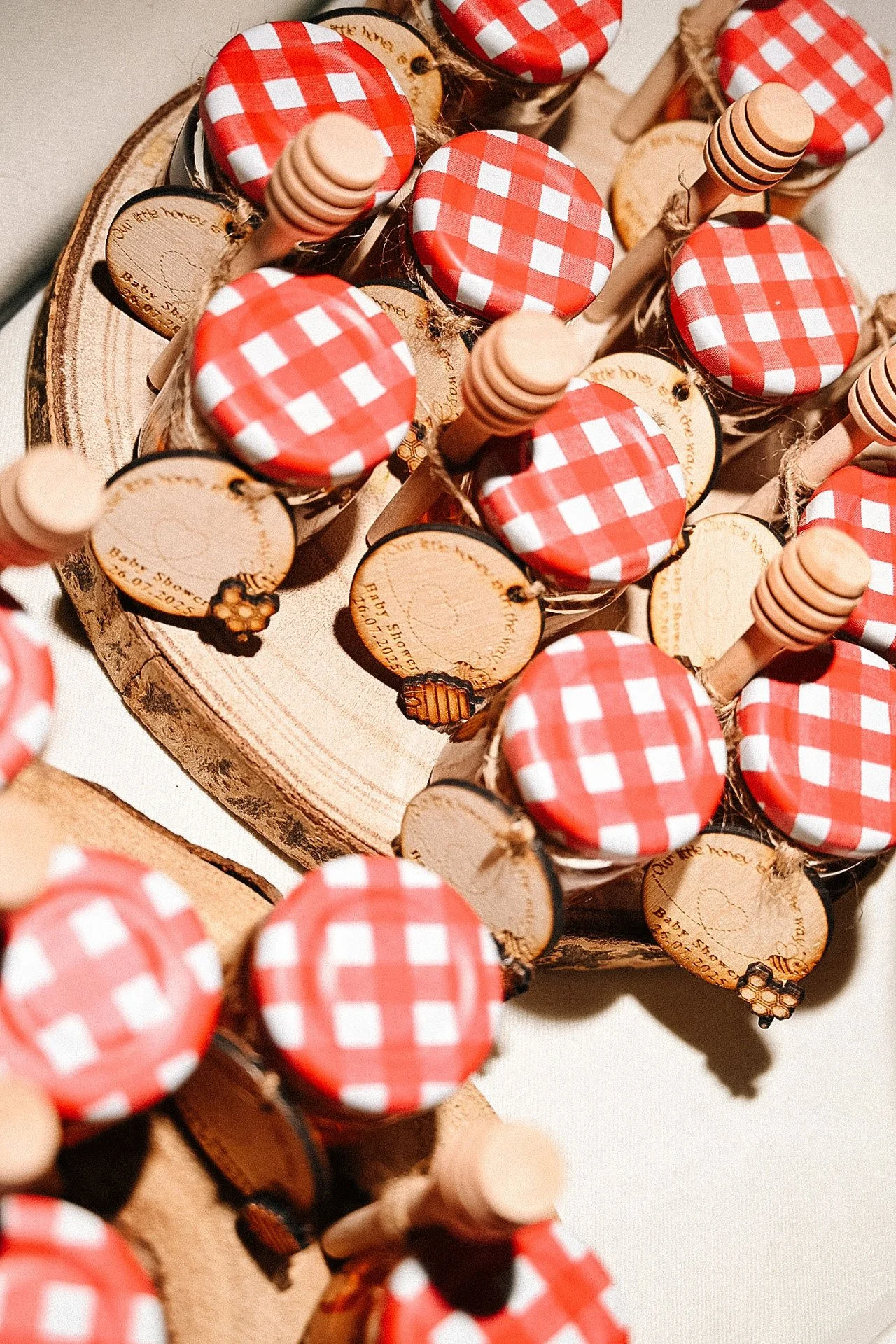 Small jars with red and white checkered lids, surrounded by round wooden tags and honey dippers, arranged on a wooden slice platter.