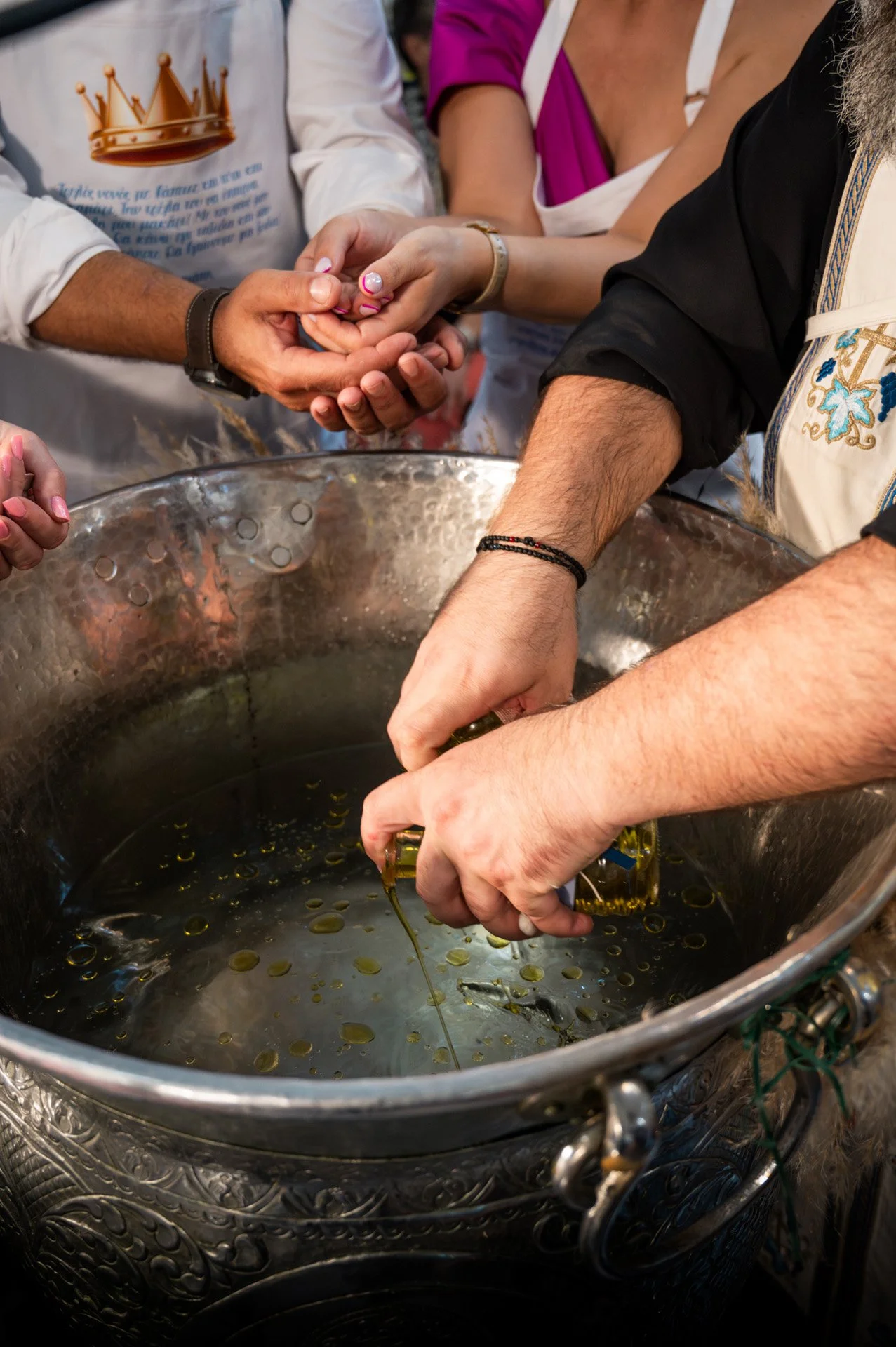 People participating in a wedding ceremony, holding hands over a large silver vessel with olive oil, during a traditional ritual.