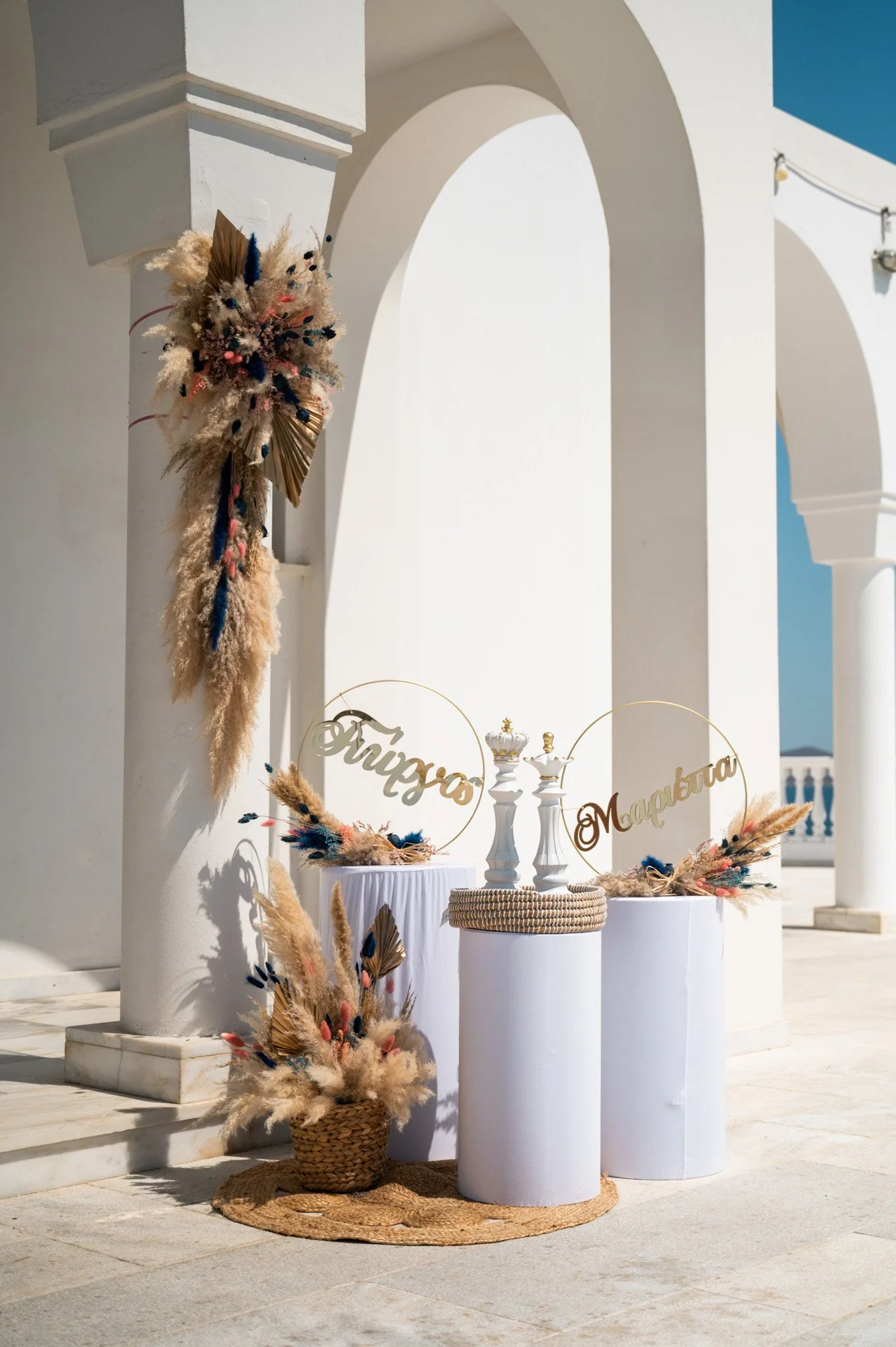 Decorative display with pampas grass, dried flowers, and gold accents on white pedestals, with signs labeled "Triuzos" and "Marelba" in a bright outdoor setting with white architectural columns and arches.