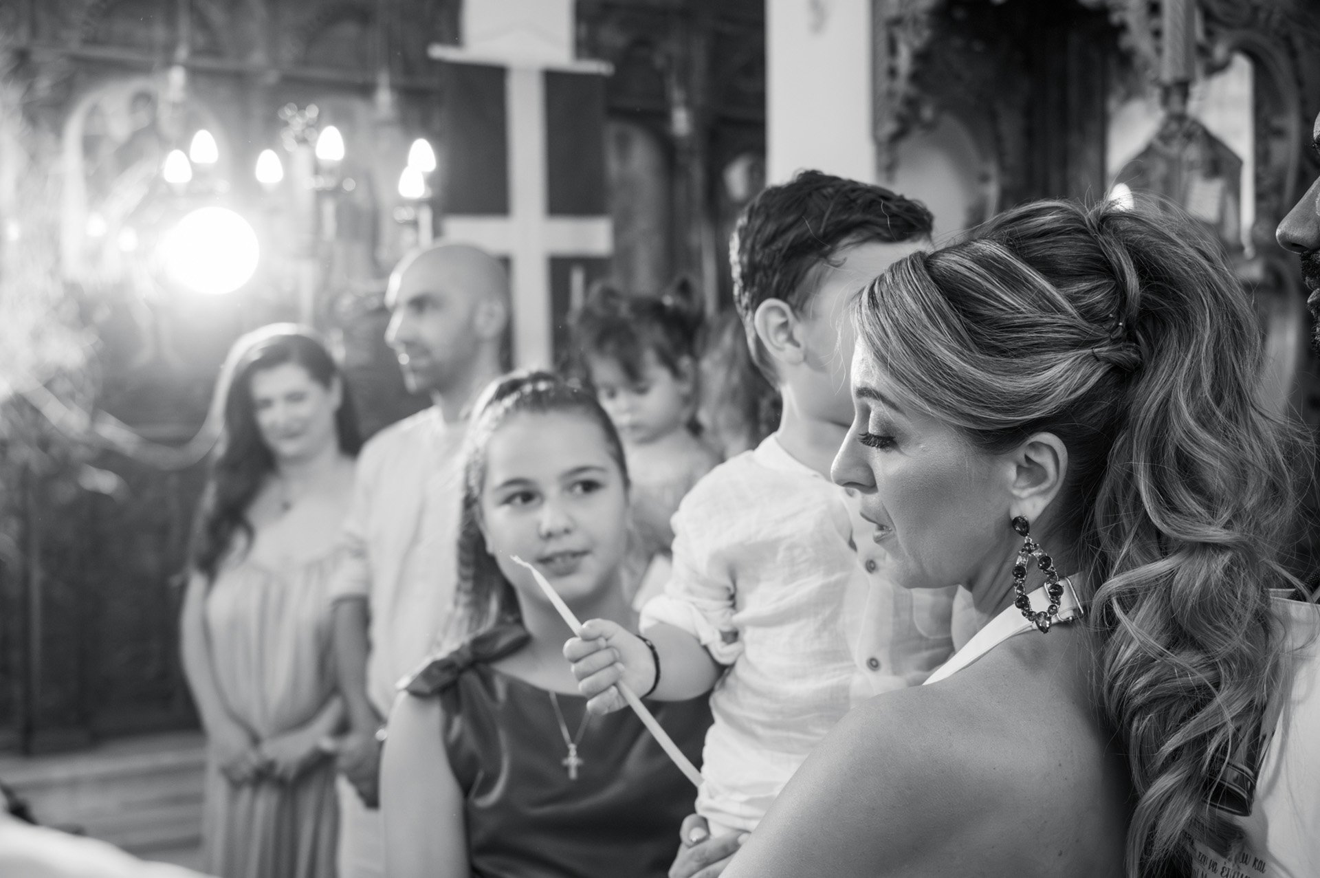 A woman with long wavy hair and earrings holds a young girl with a candle, surrounded by several people in a church with religious decorations and cross in the background.
