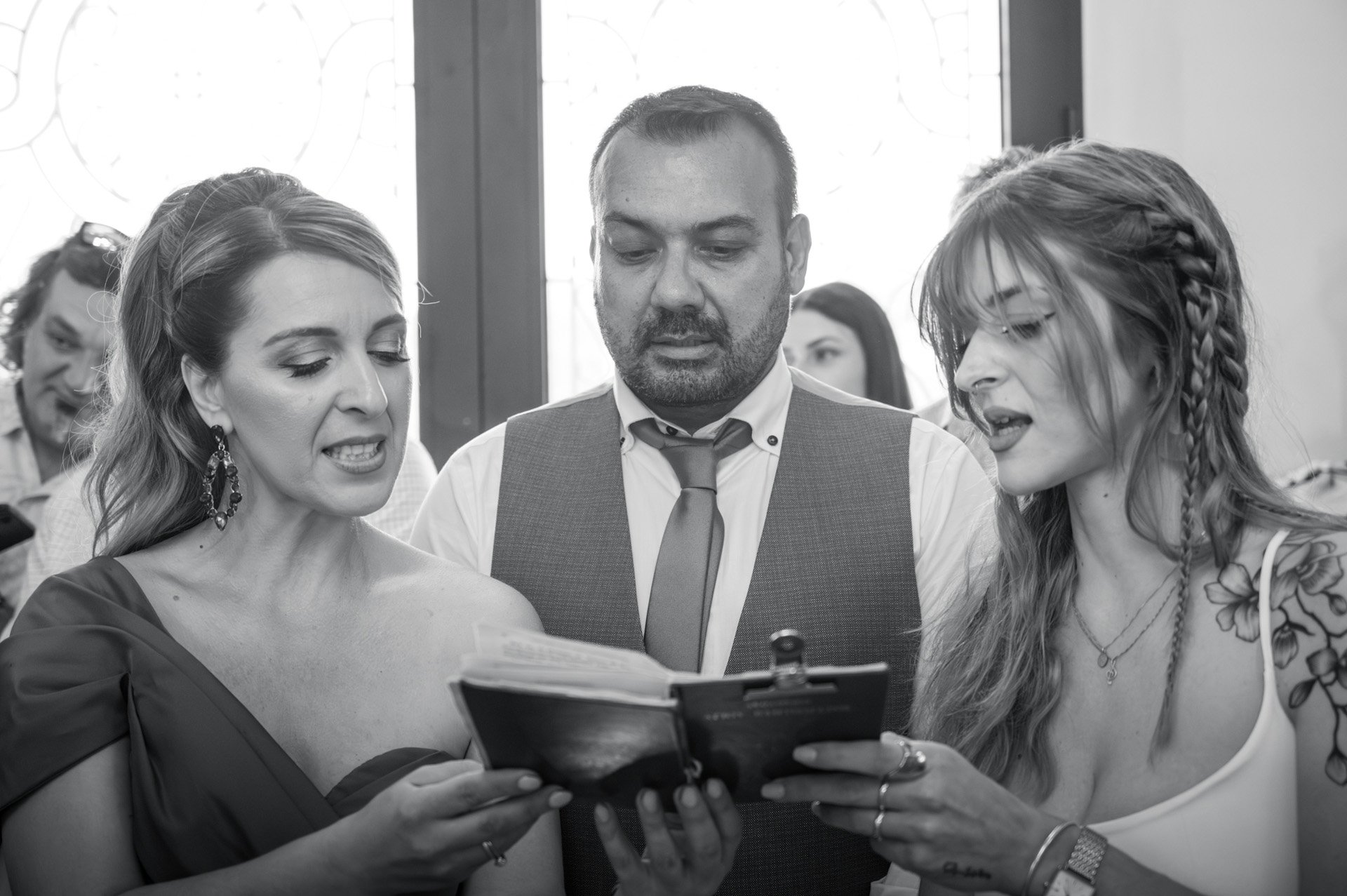 Three people looking at a small booklet or photo album closely, with two women on either side and a man in the middle, in a busy indoor setting.