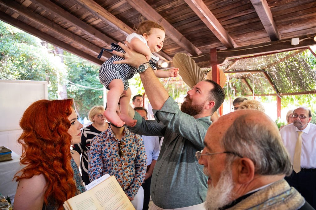 A man lifting a smiling baby girl in the air during a gathering, with several onlookers including a woman with red hair holding a book. The scene is outdoors with a wooden ceiling and natural light.
