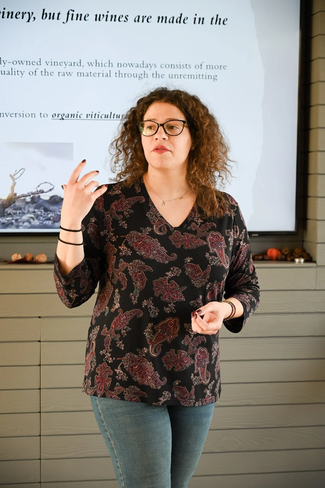 A woman with curly brown hair, glasses, and wearing a black and red paisley patterned top, standing in front of a presentation screen during a talk.
