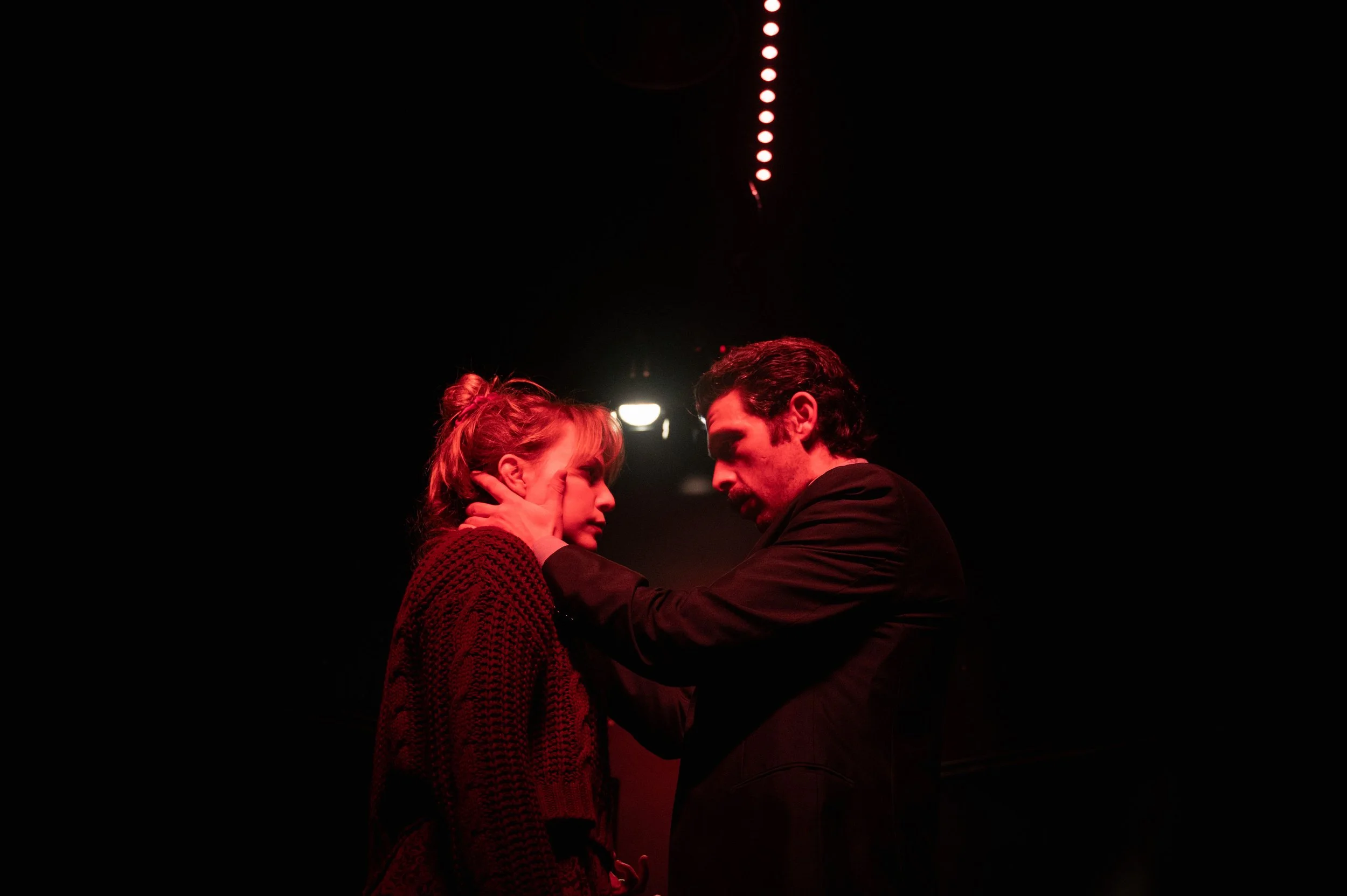 A man holding a woman's face in his hands, both with serious expressions, in a dark setting with red lighting.