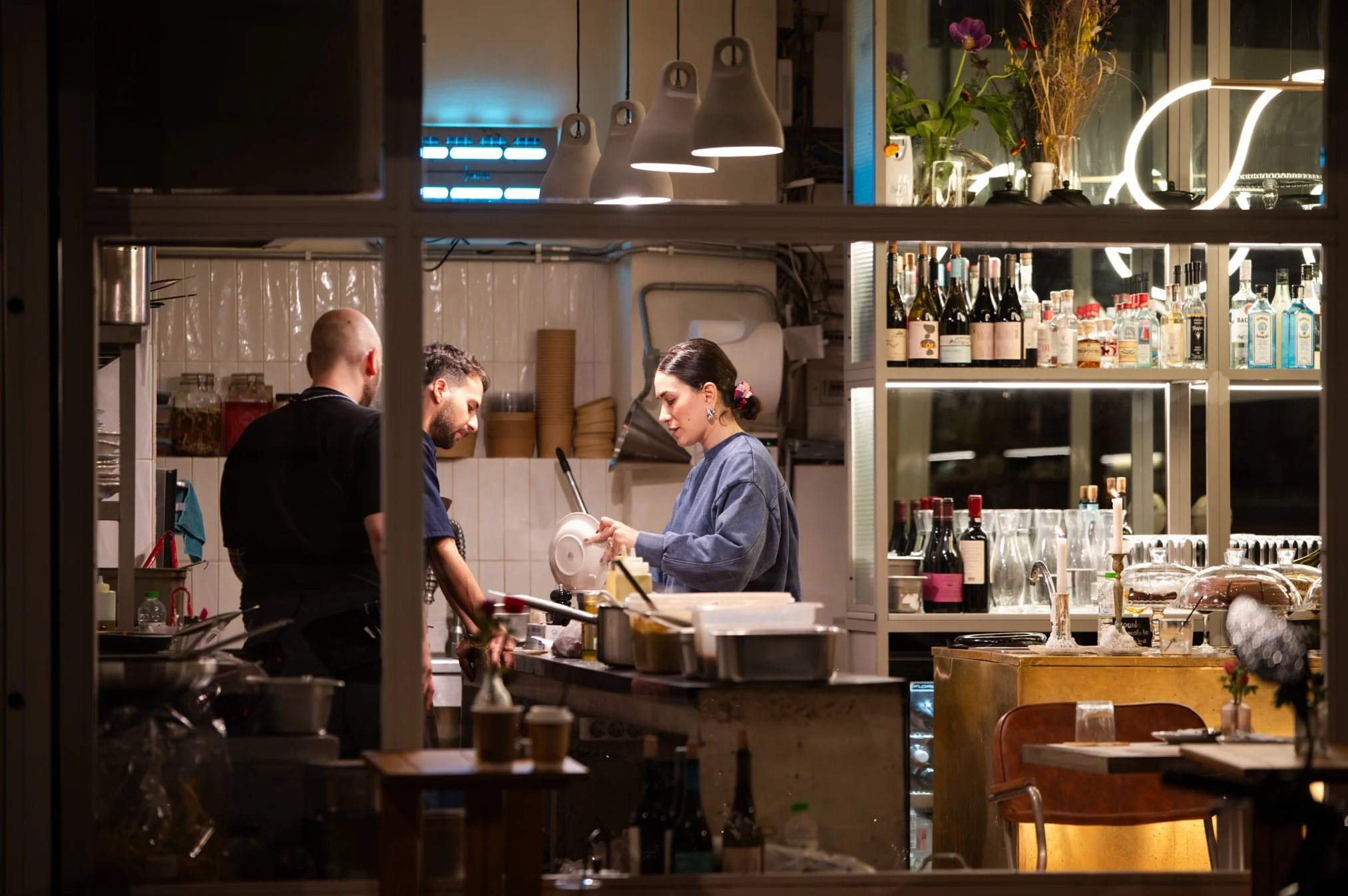 Cook and waitress working behind the bar at a restaurant or cafe, with shelves of wine and liquor bottles, flowers, and kitchen utensils visible in the background.