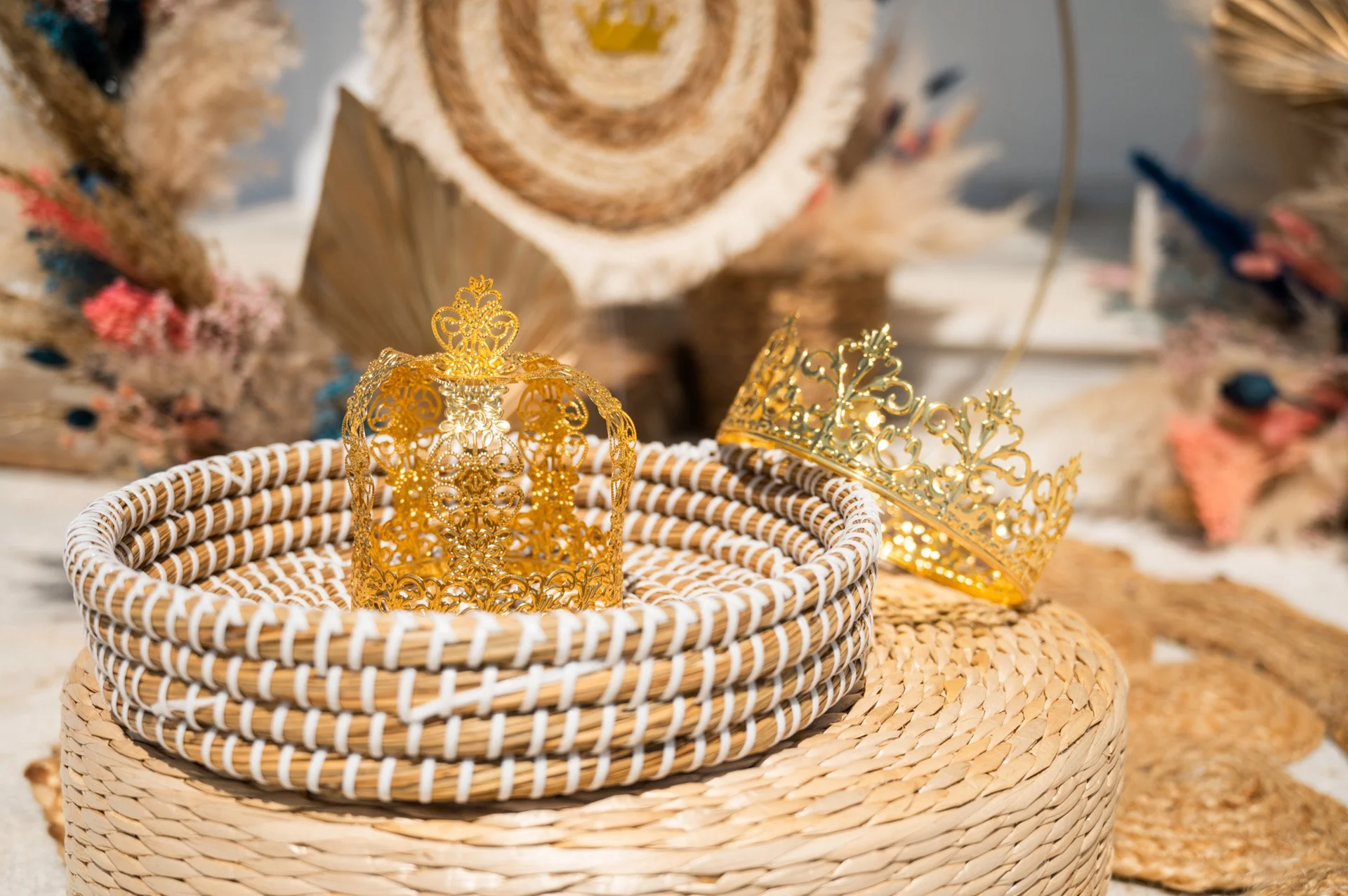 Two gold crowns placed on woven basket and a woven round surface, with decorative dried flowers and wooden pieces in the background.