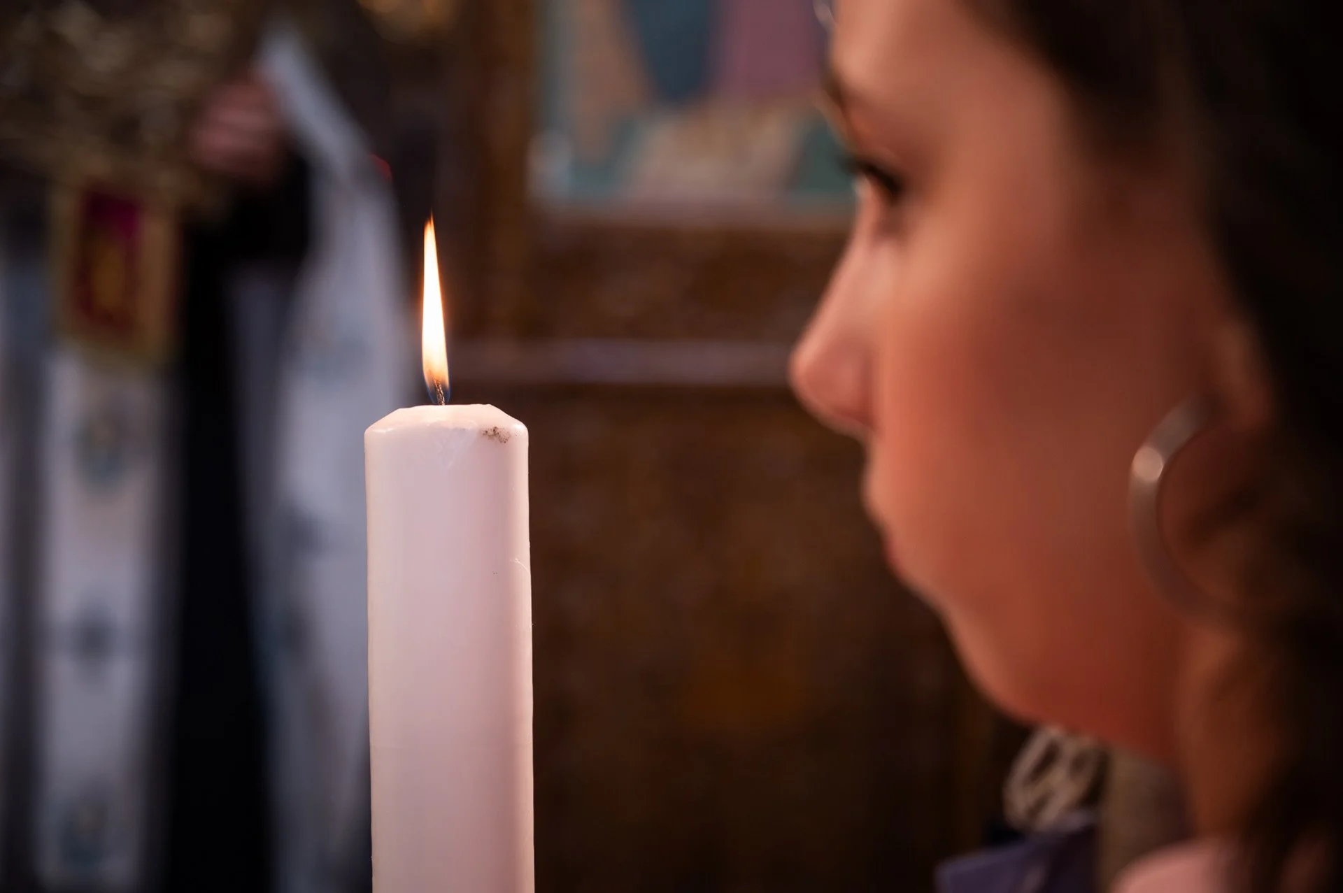 A woman with dark hair and hoop earrings is seen in profile, looking at a lit white candle.