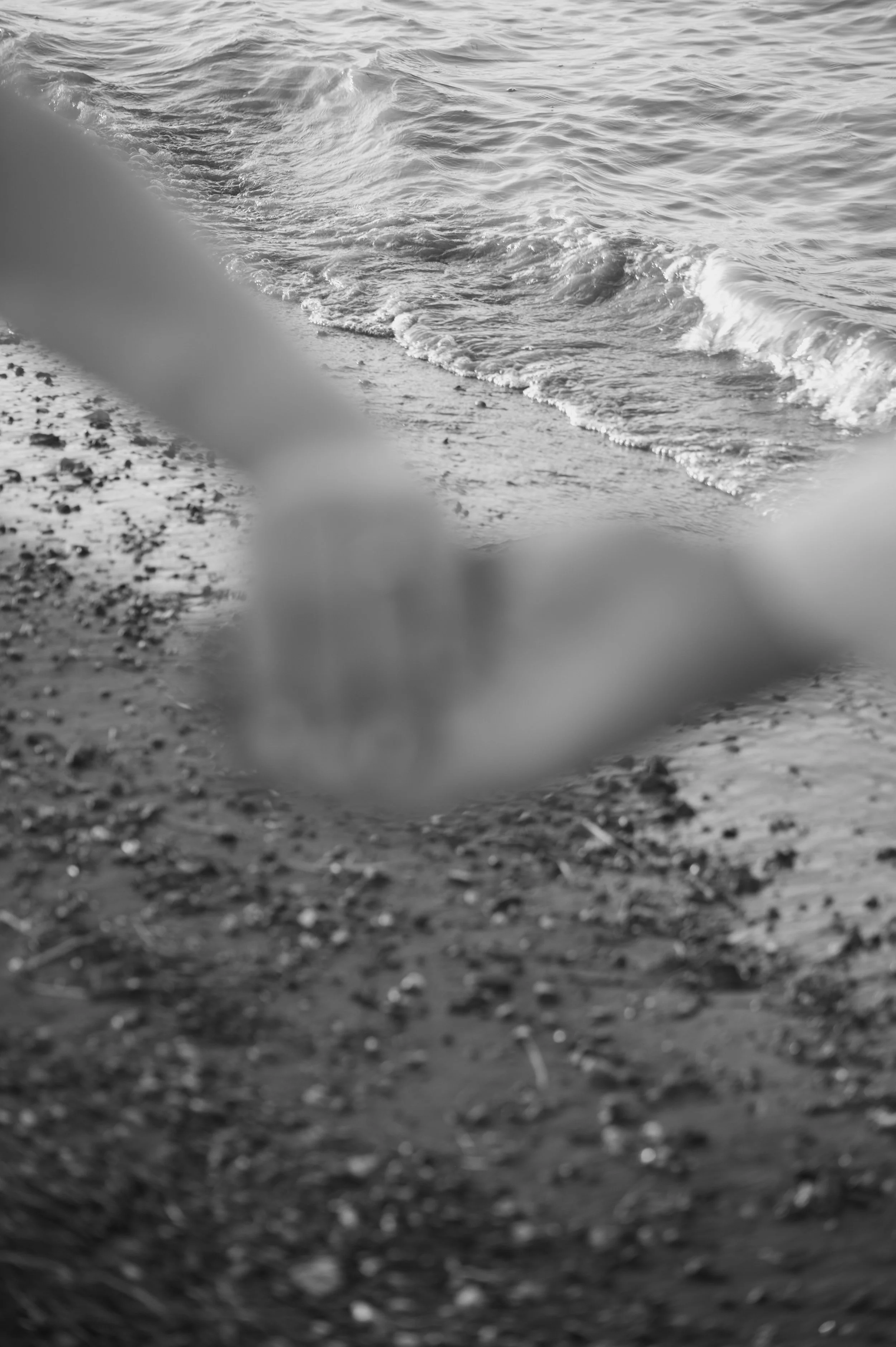 Blurry black and white photo of a beach with granular sand and small stones, and water with waves in the background.