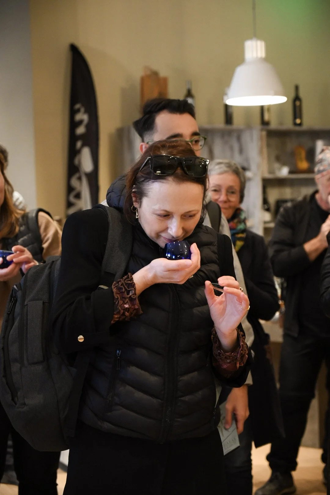 A woman smelling a glass of wine at a social gathering indoors, surrounded by other people.
