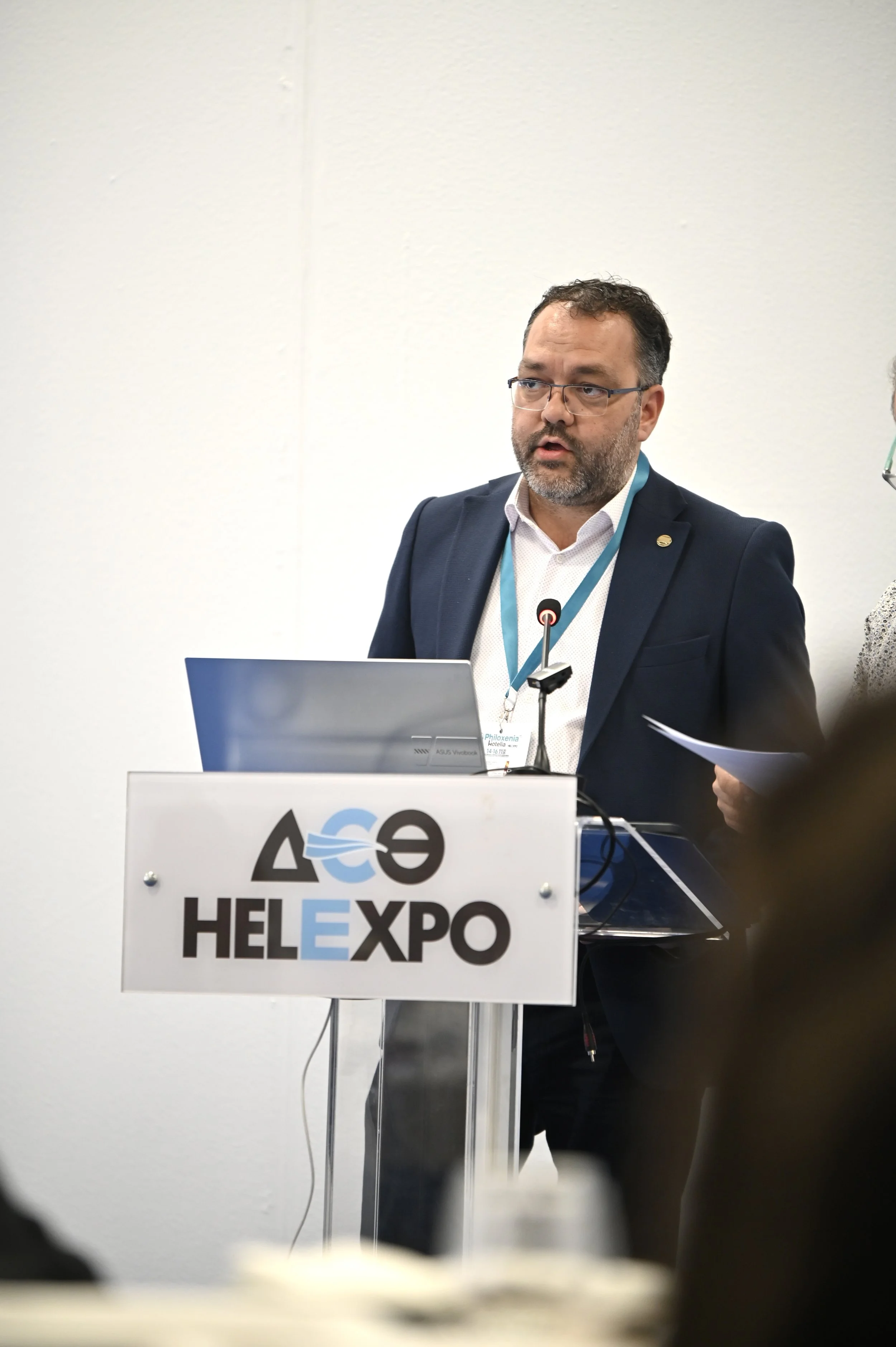 A man in a suit giving a presentation at a conference with a sign that says 'HELLEXPO' on the podium.