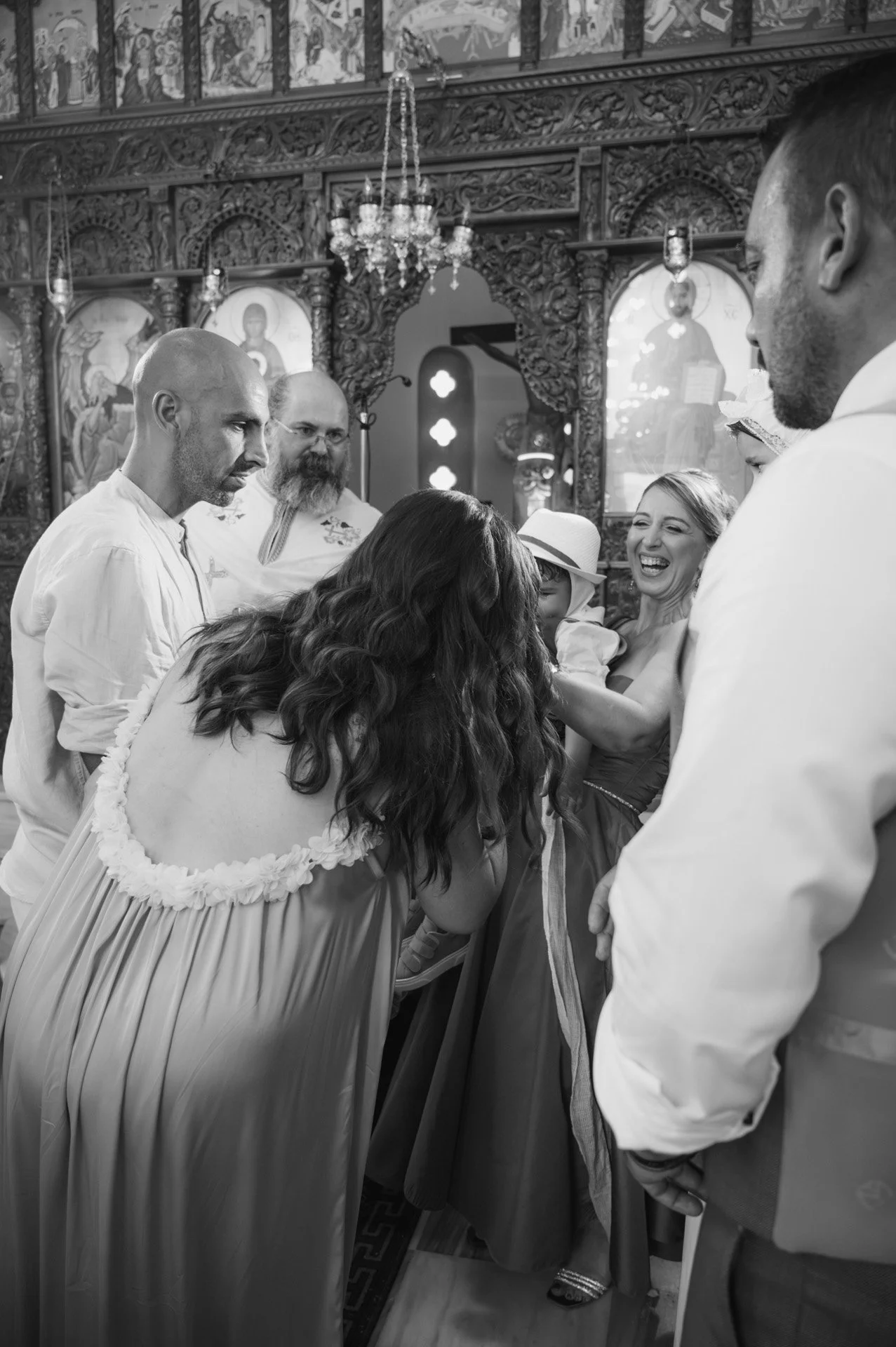 A group of people dressed in formal attire inside an ornate church with religious icons and chandeliers, participating in a religious ceremony or gathering.