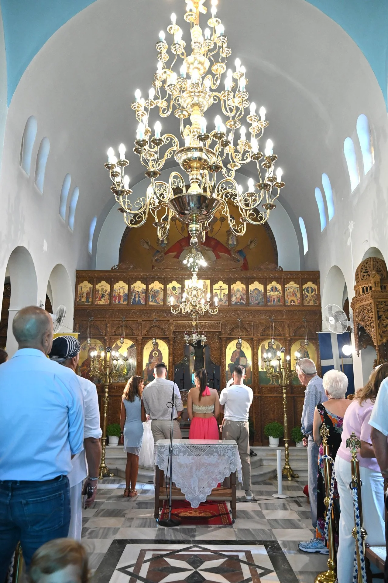 Inside a church during a wedding ceremony, with guests standing and facing the altar, a large chandelier hanging from the ceiling, wood-carved iconostasis behind the altar, and religious icons depicting saints and biblical scenes.