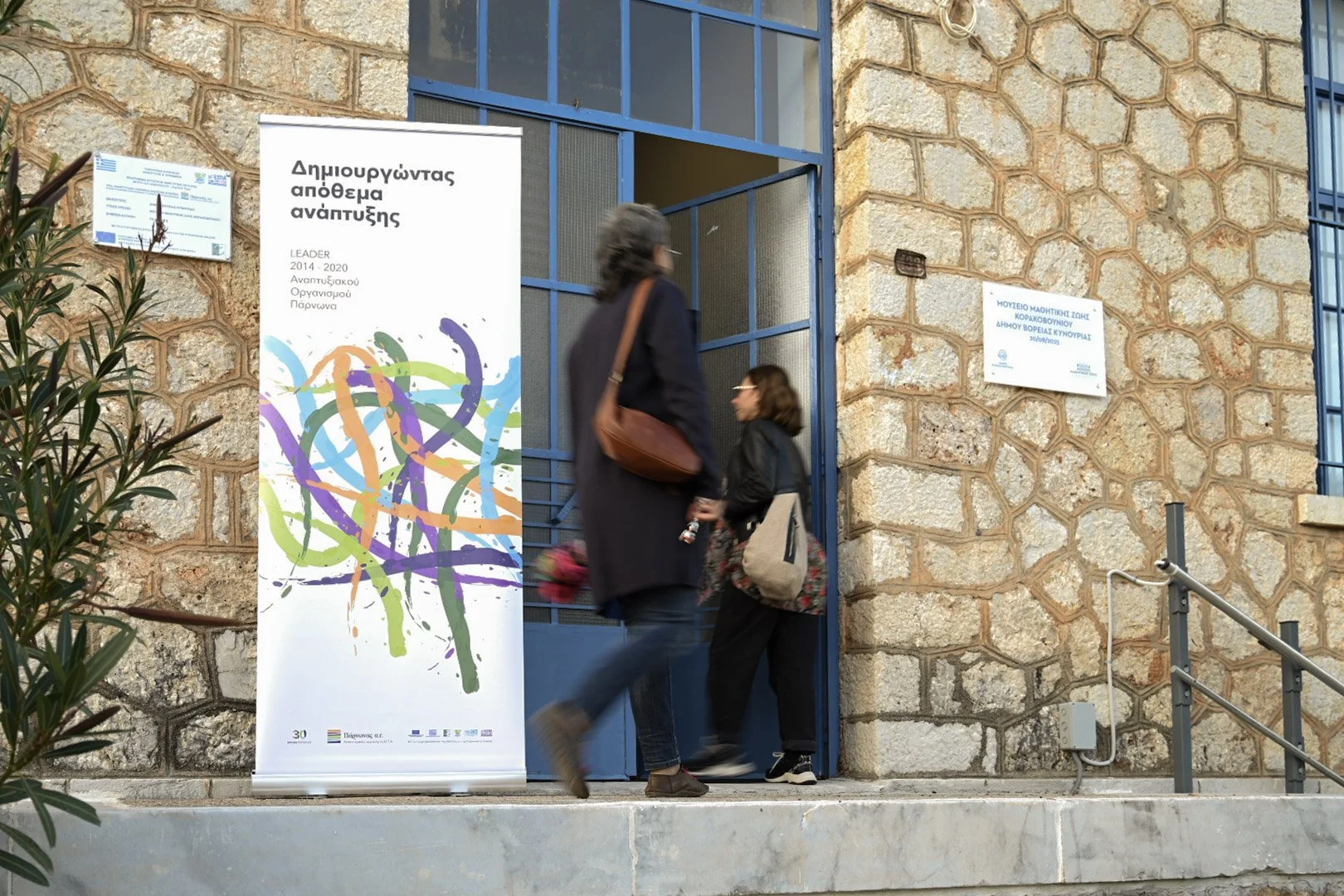 People entering a building with stone wall exterior, a blue door, and posters on the wall. One person is passing through the door while another stands nearby.