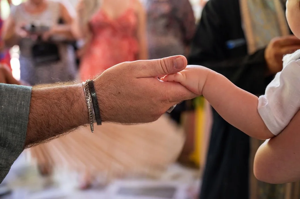 A close-up of a man with a hairy arm and bracelets holding the hand of a young girl with light skin in a white shirt, against a blurred indoor background with people.