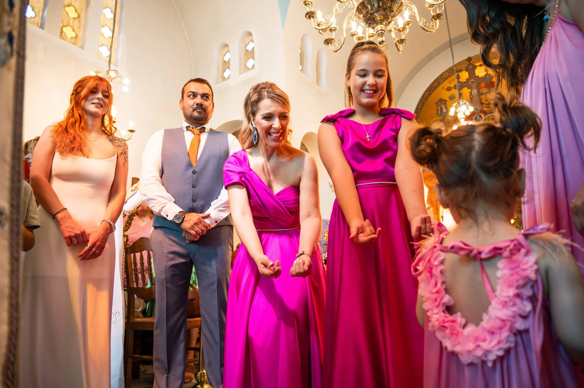 A group of people, including women and children, gathered in a church during a celebration, possibly a baptism or wedding, with some dressed in pink dresses, in a church with high ceilings and ornate lighting fixtures.