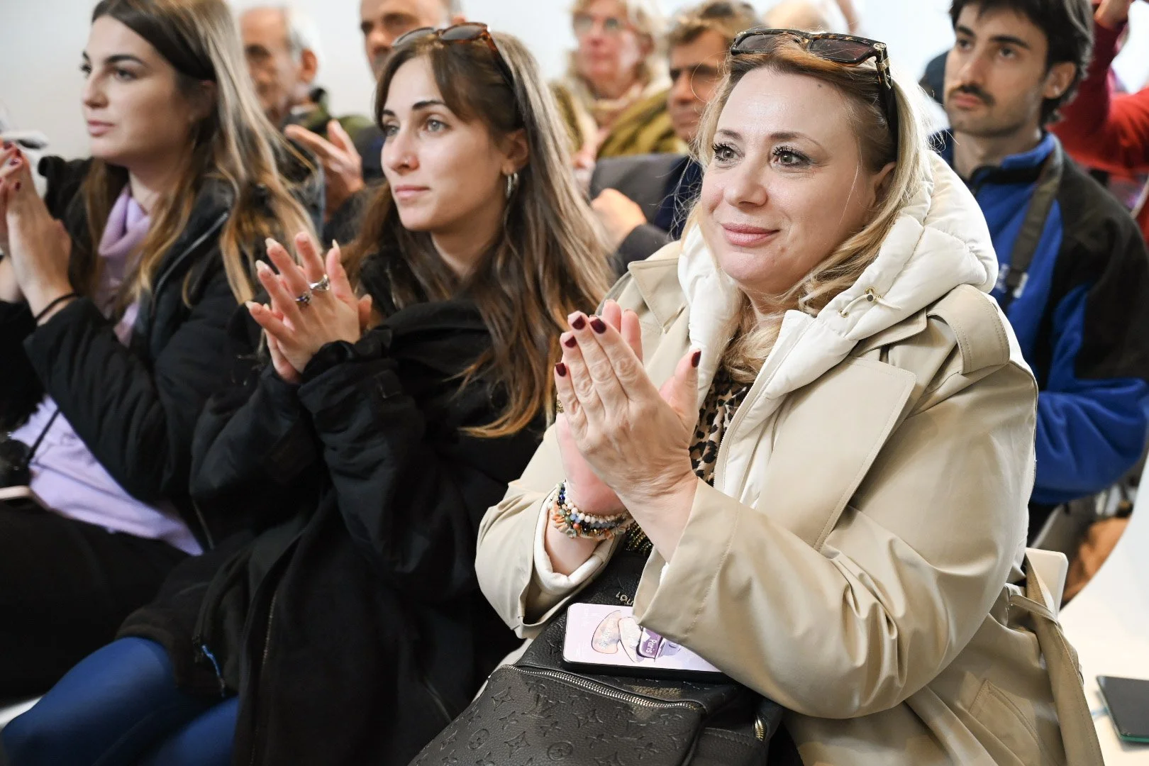 A group of people sitting and clapping at an indoor event, including a blonde woman in a beige jacket in the foreground.