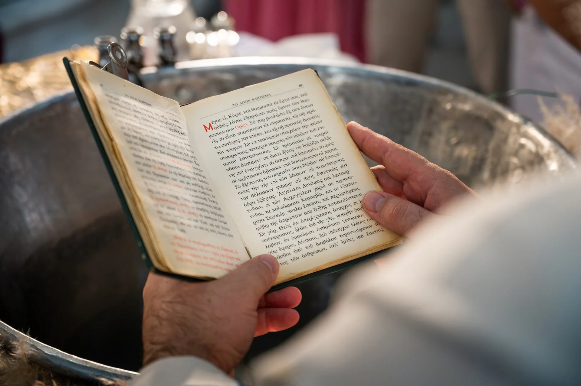 A person holding an open book with Greek text while resting their hands on a metal basin or sink.