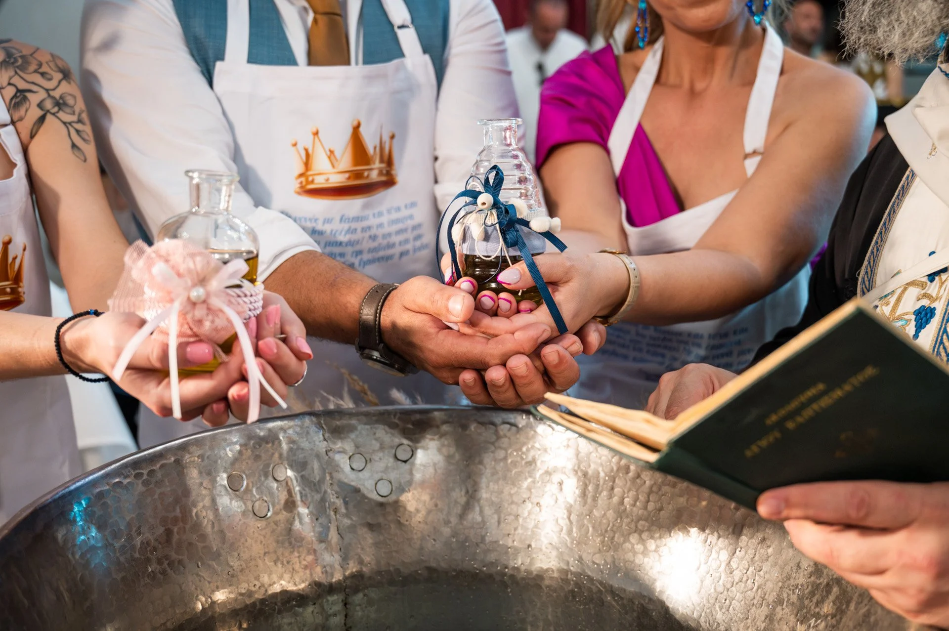 People participating in a ceremonial event, holding bottles with ribbons over a large metallic basin, with an individual reading from a book in the bottom right corner.