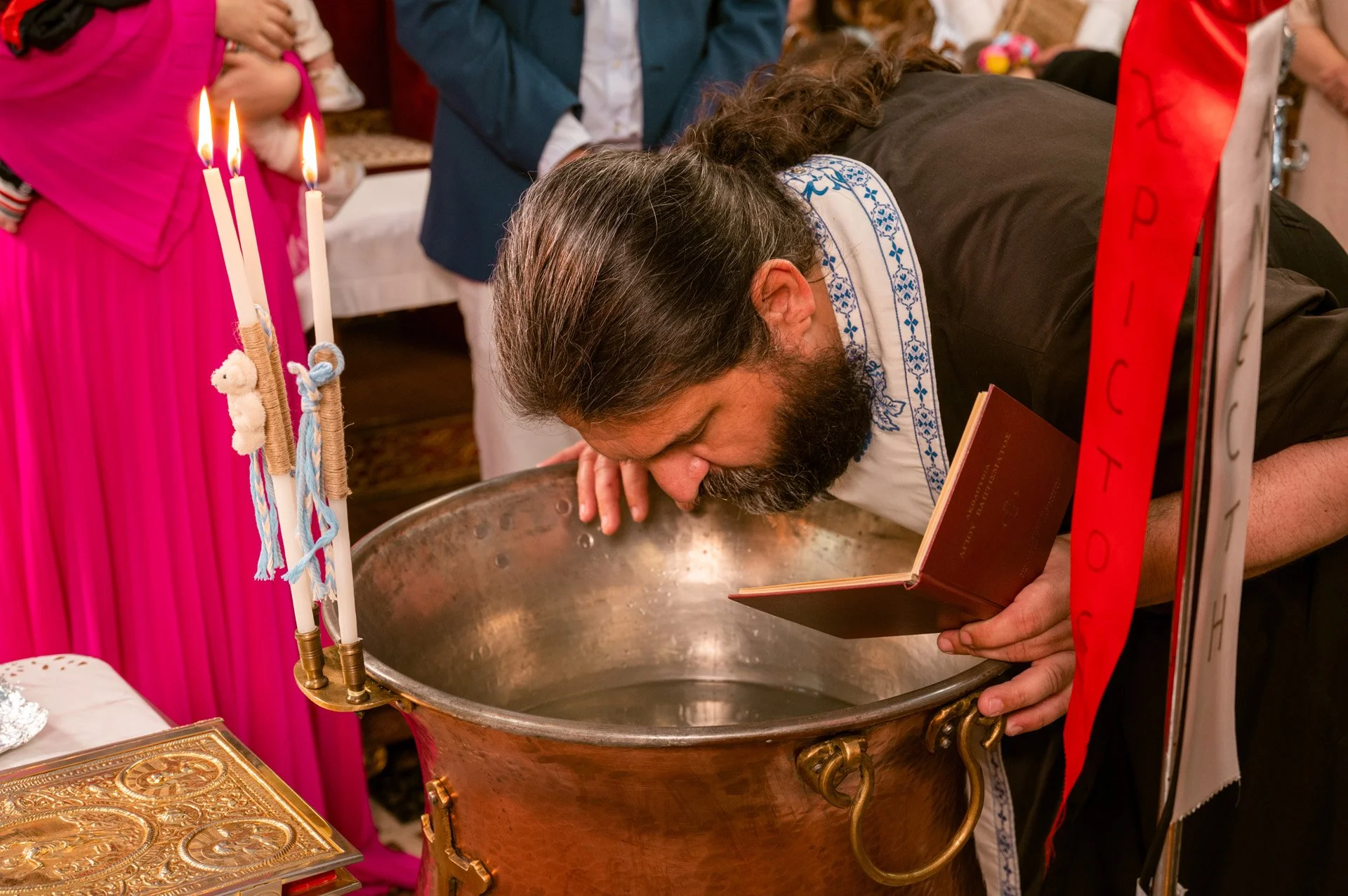 A man with long hair and a beard is kissing a baptismal font inside a church while holding a book. There are candles nearby, and people in formal attire are in the background.