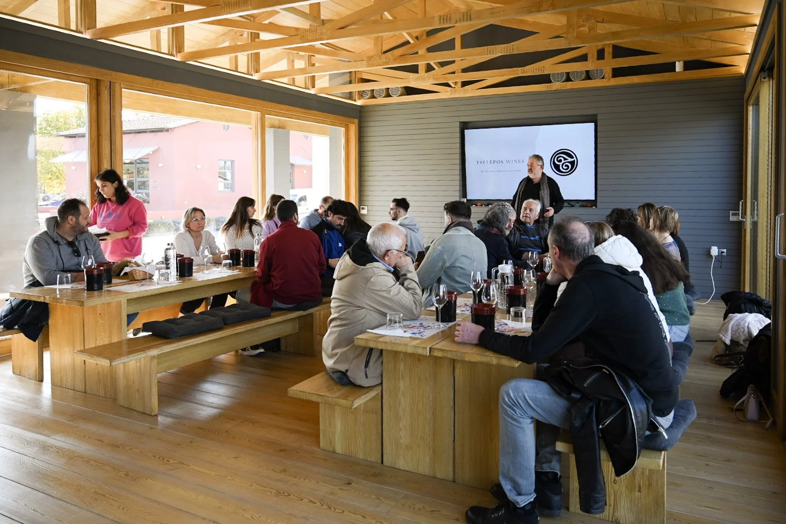 A group of people attending a presentation or tasting event in a modern wooden interior with large windows, seated around tables with glasses of wine, while a presenter speaks in front of a screen displaying the logo 'Iselepos Wines'.