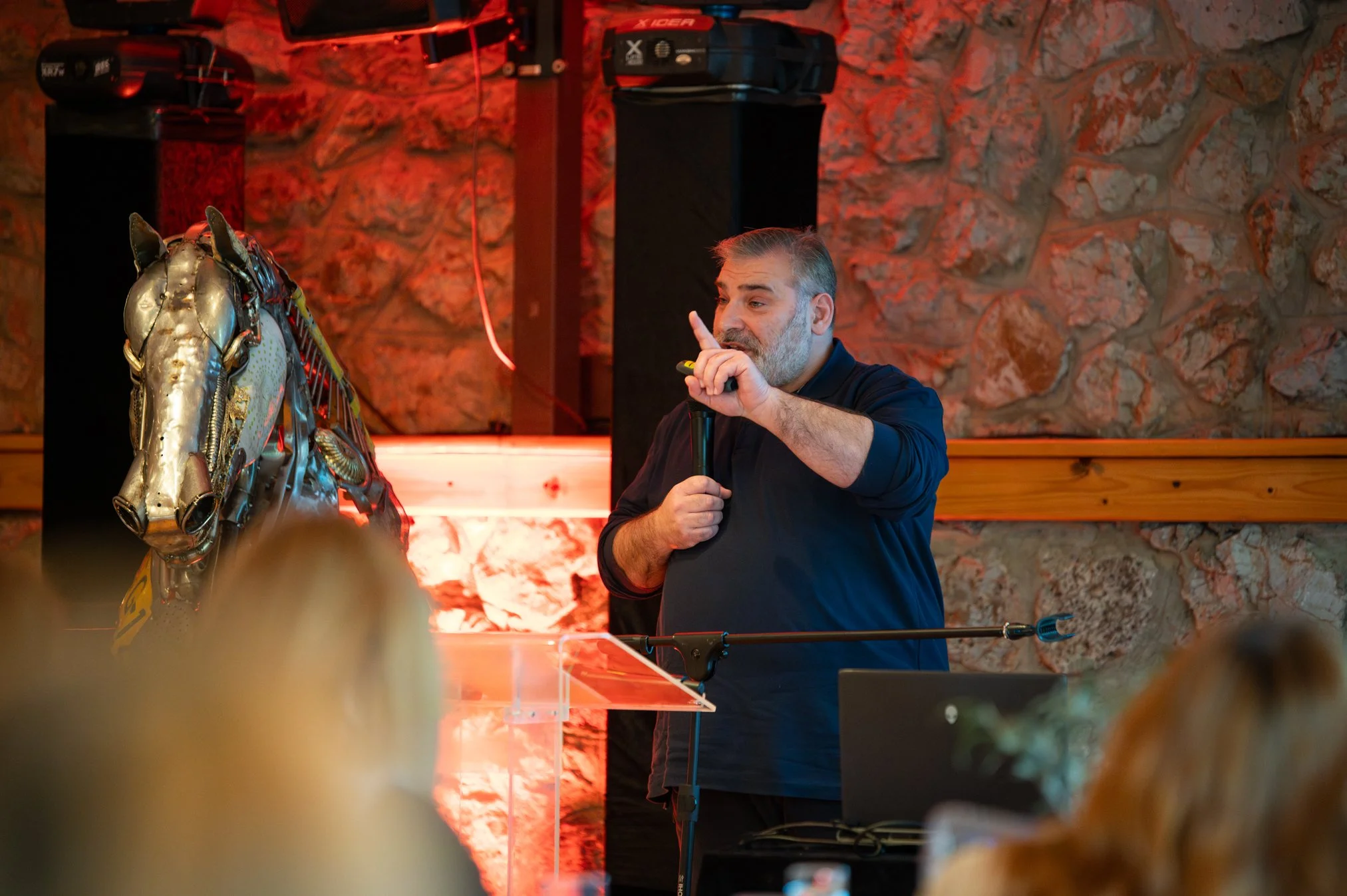 A man giving a presentation indoors, with stone walls and red lighting, near a decorative horse sculpture.