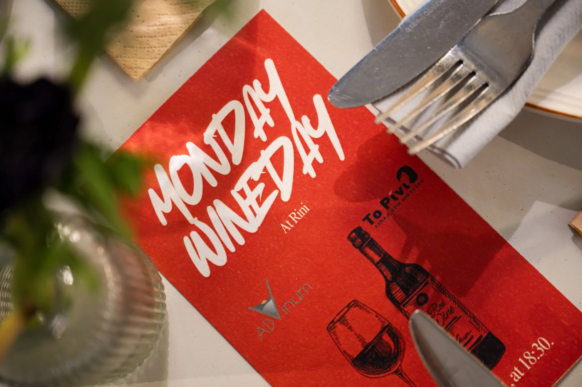 Red advertisement card for Monday Wine Day at Rimi with illustrations of a wine bottle and glass, located on a table with silverware and napkins.
