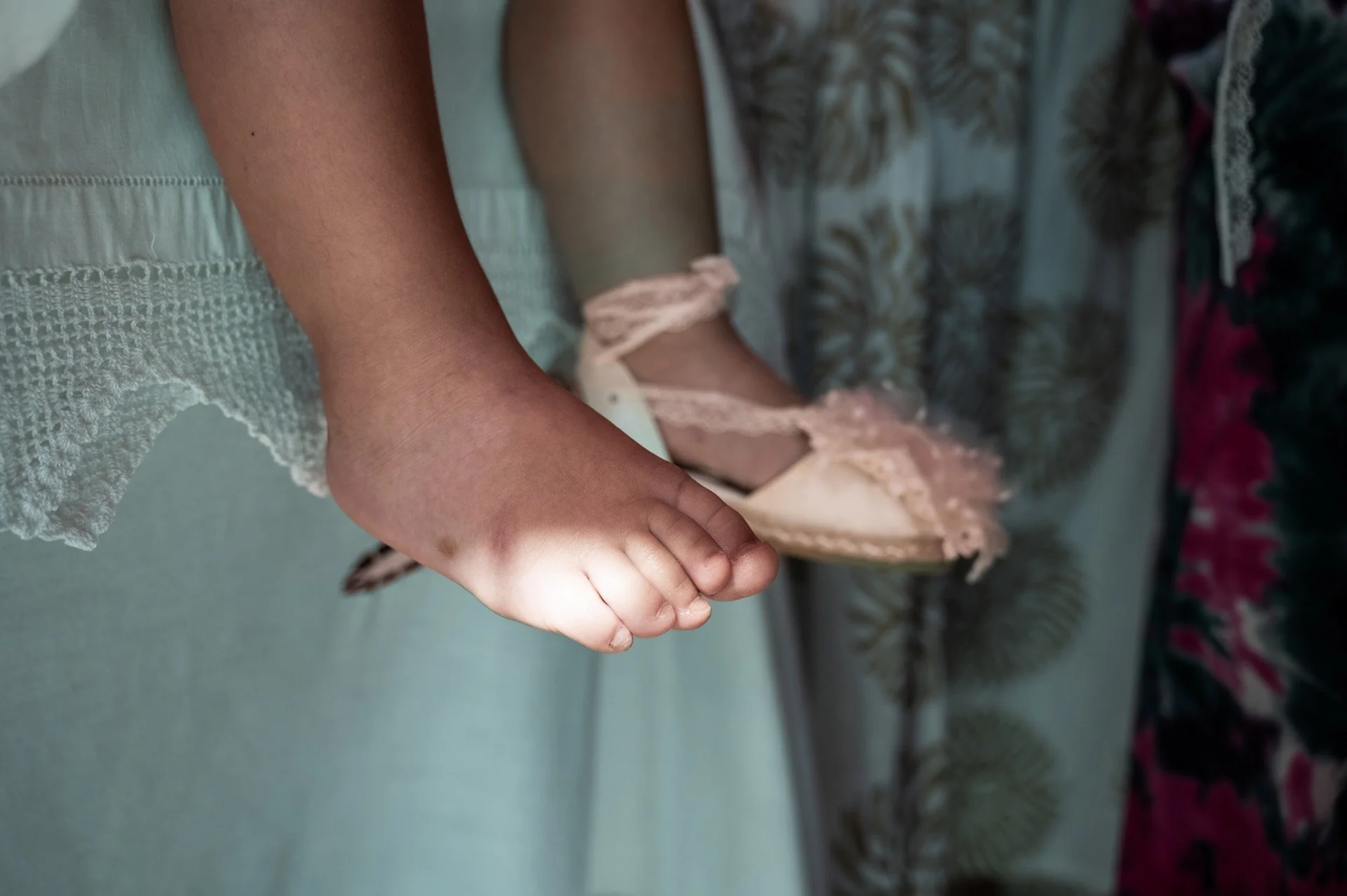 Close-up of a baby’s foot with toes, resting on a surface, with a part of the baby's leg and a lace dress visible. The background includes patterned fabric and floral decorations.