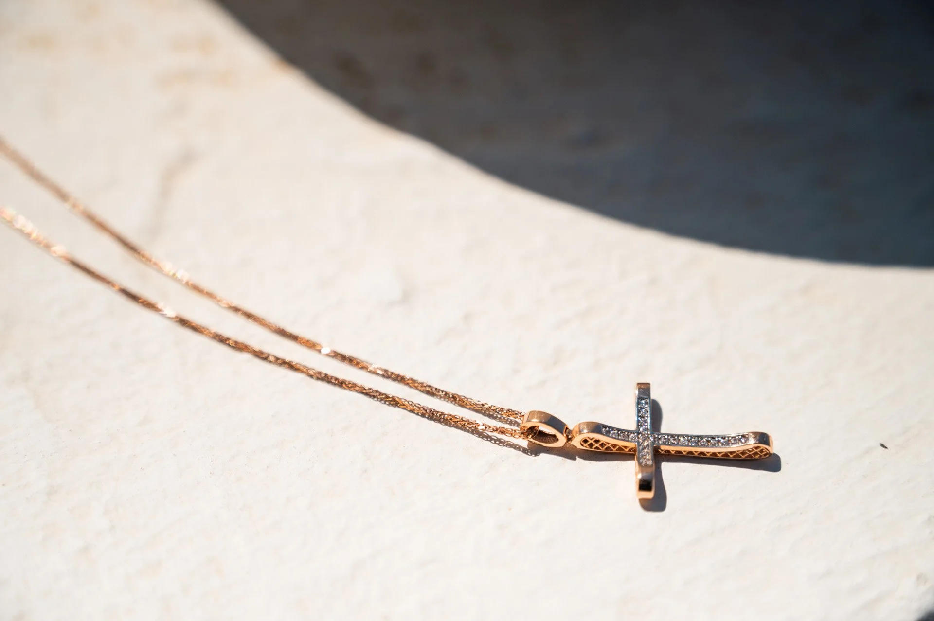 A rose gold necklace with a cross pendant adorned with small diamonds resting on a beige surface with shadows.