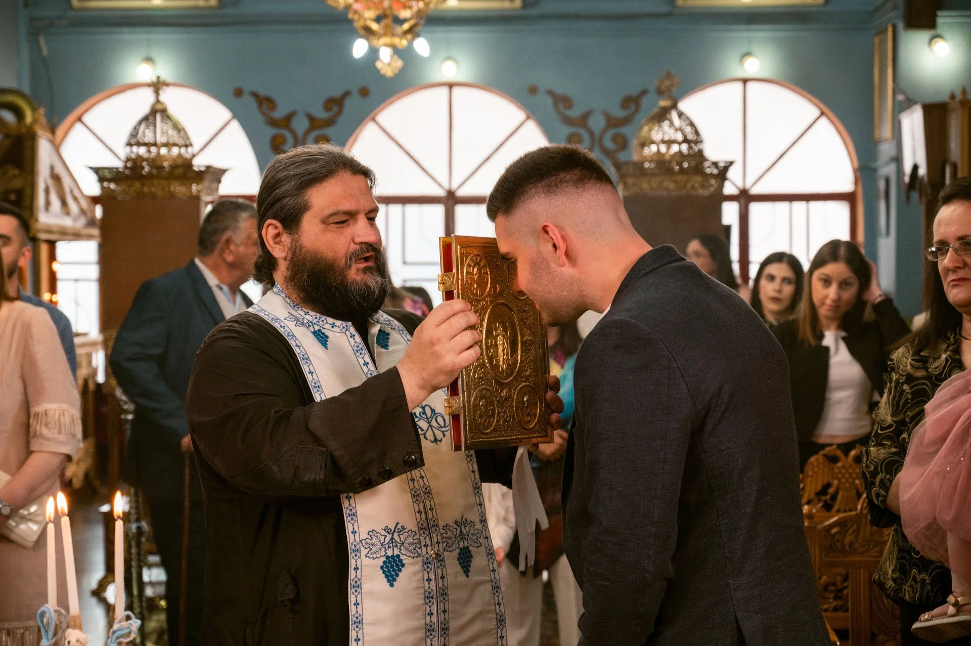 A priest in religious attire holding an ornate religious book, blessing a man with his forehead in a church setting during a ceremony, with several onlookers in the background.
