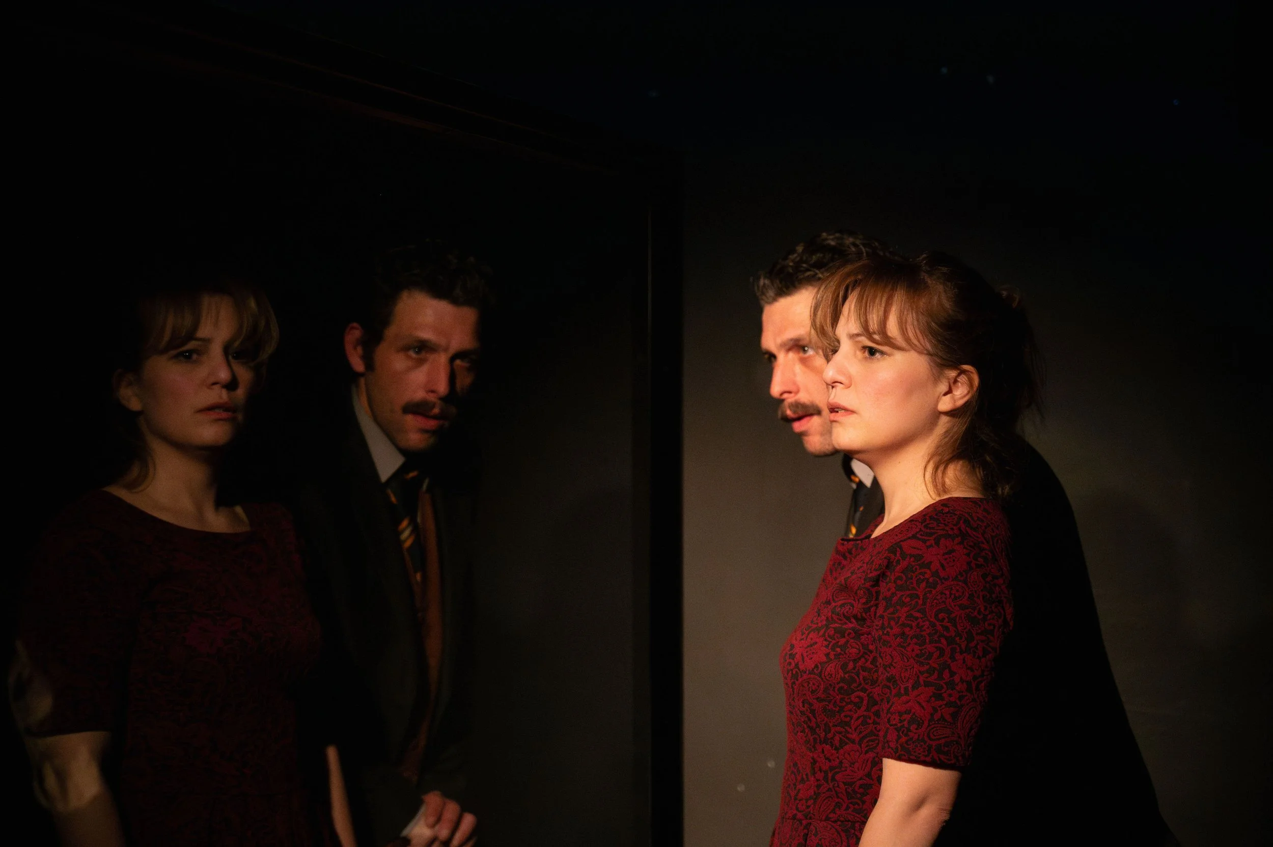 Four actors standing in the dark, looking thoughtful, with a mirror reflecting their images.