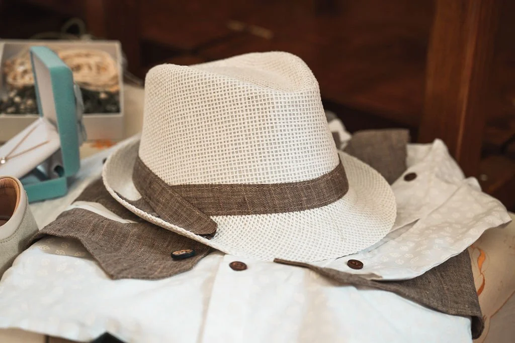 A white straw fedora hat with a brown band resting on a partially opened gray and white shirt. In the background, there are jewelry boxes and other items, all on a cluttered surface.