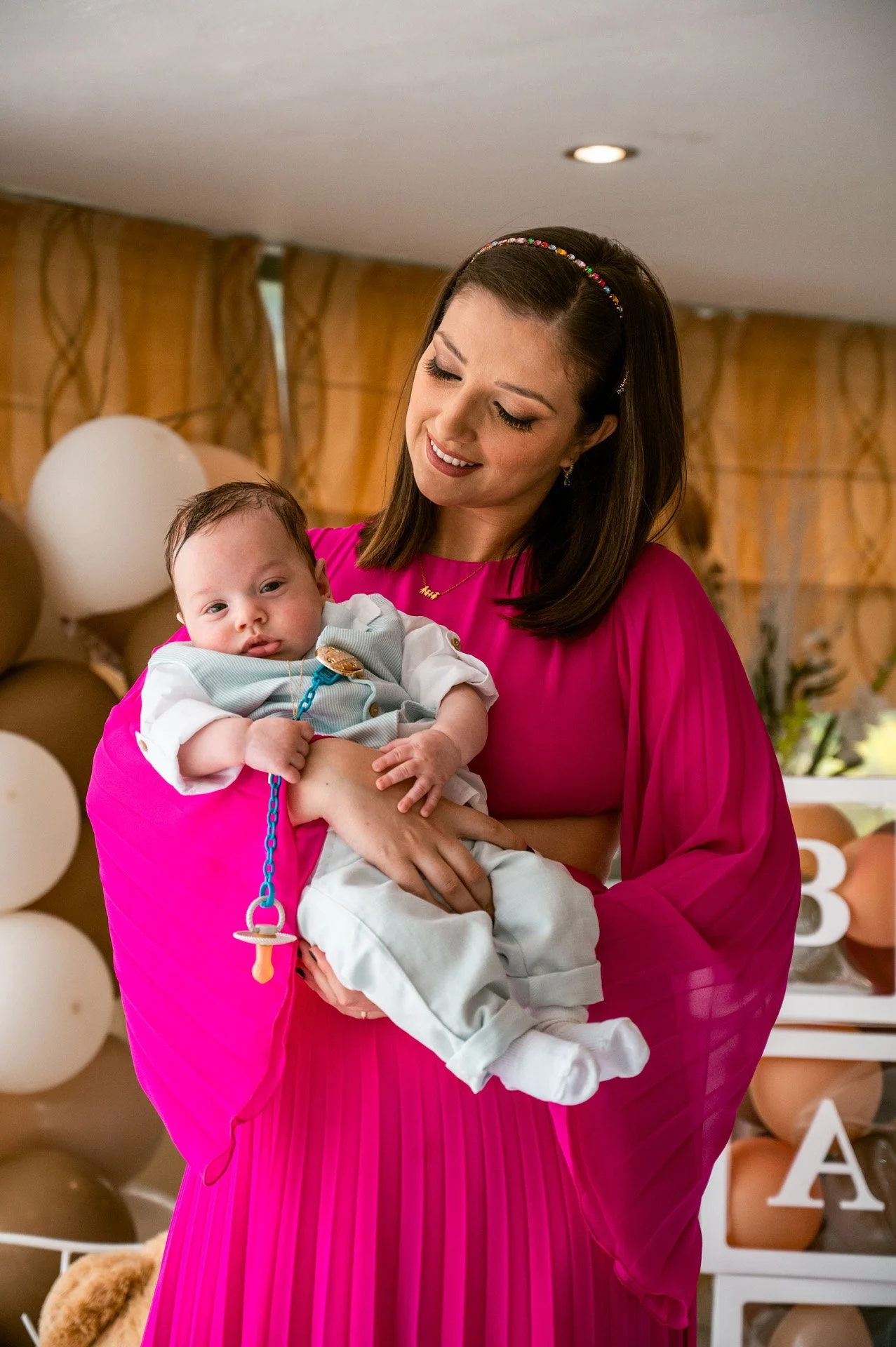 A woman in a bright pink dress holding a baby in a light-colored outfit with a pacifier clip, smiling at the baby indoors with balloons and decorations in the background.