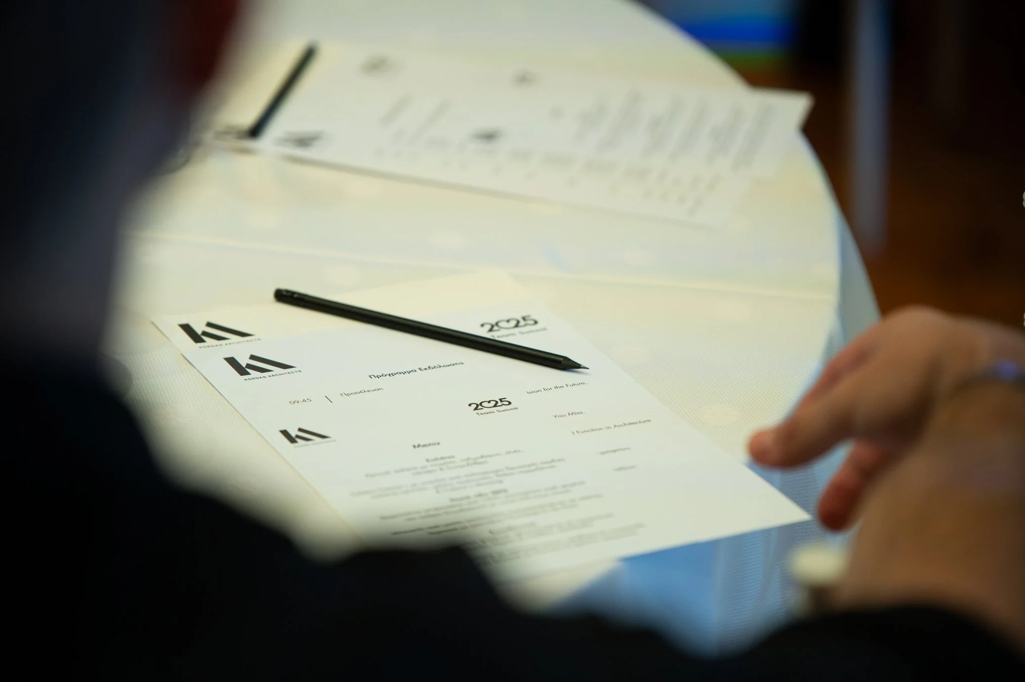 A printed schedule or program for an event titled 'Protoyping Exhibition' with the subtitle 'Vision for the Future', including a time 09:45 and room 'Room 225'. A black pen rests on top, and hands are visible at the bottom right, pointing at the docu