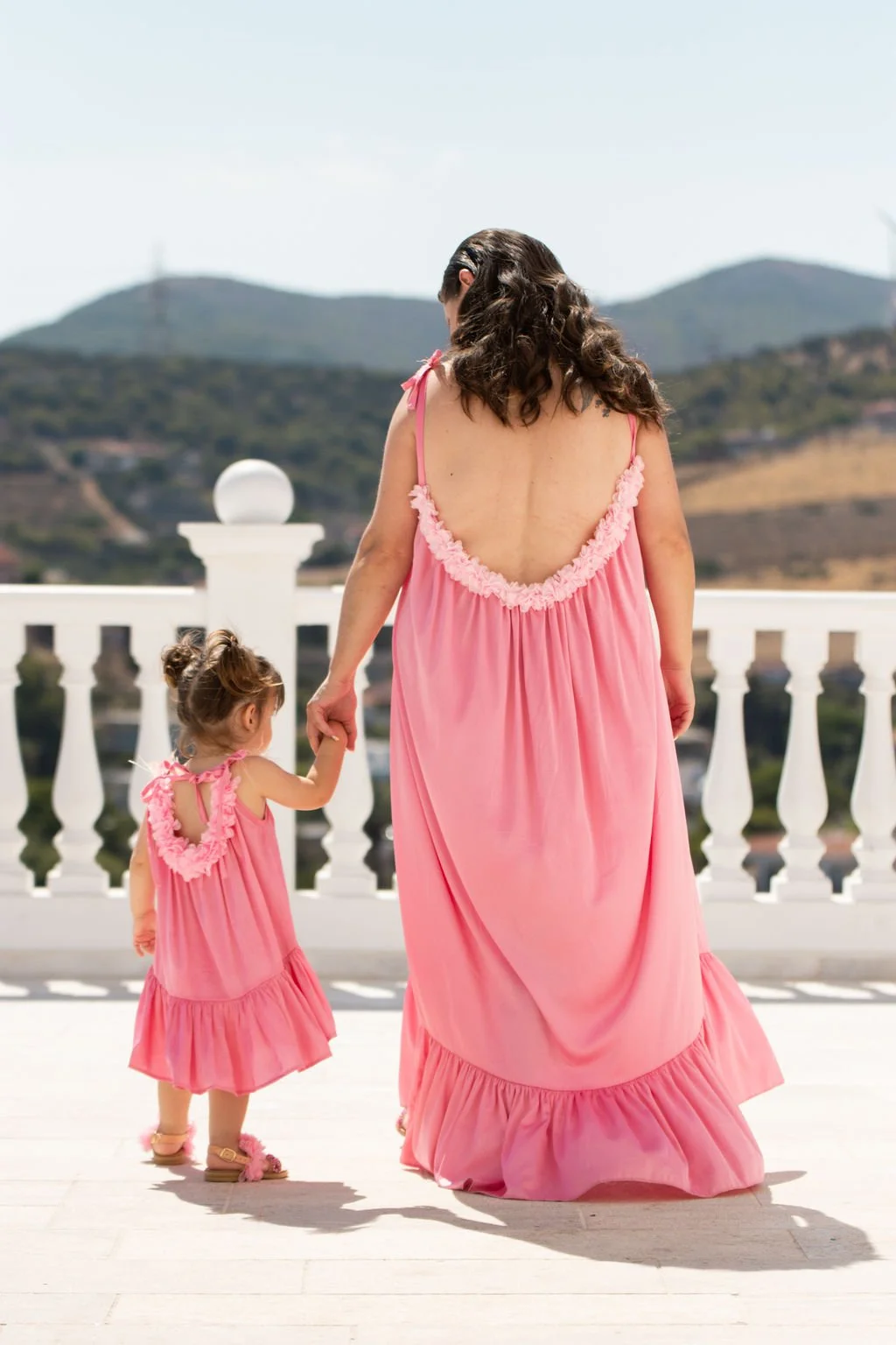 A woman and a young girl, both dressed in matching pink dresses, walking hand in hand on a balcony with a scenic hilly landscape in the background.