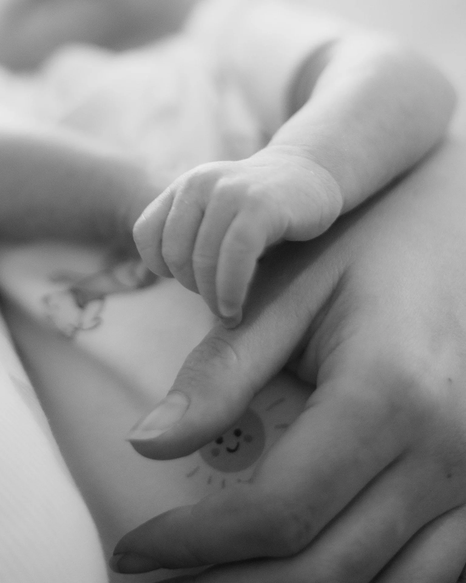 Close-up of a baby's tiny hand holding a woman's finger, with a small smiling sun tattoo visible on the woman's hand.