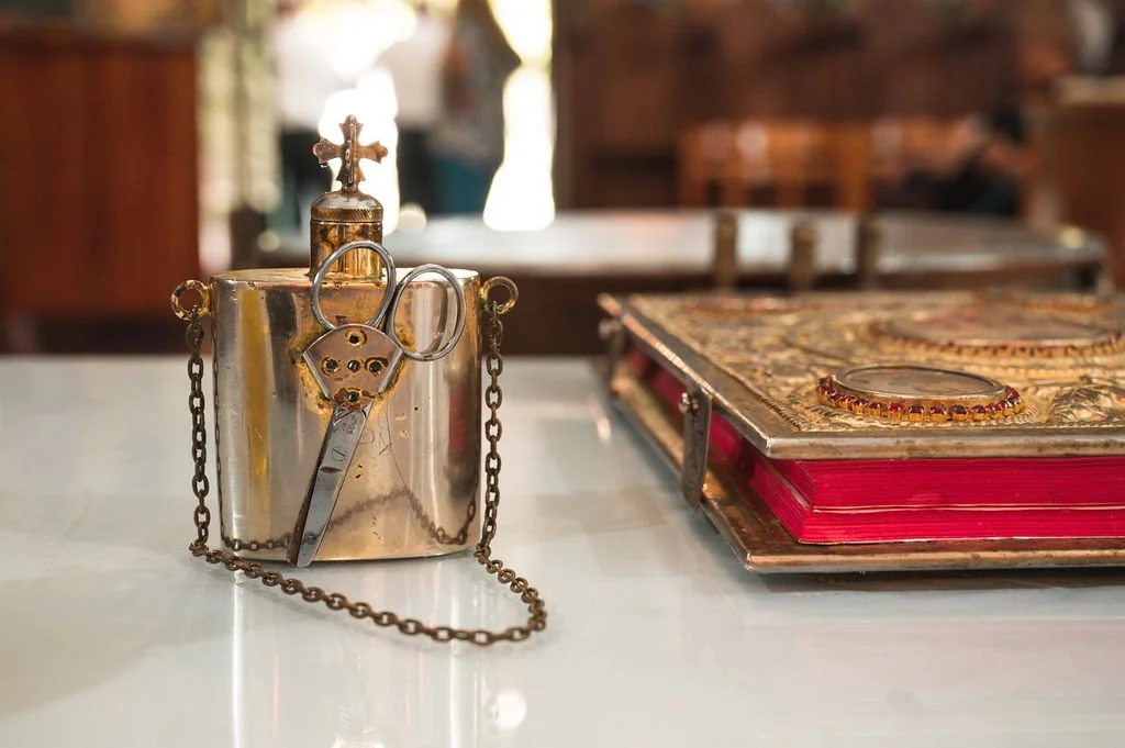 A small, shiny silver religious container with a cross on top, chains on the sides, and a ring and scissors hanging from it. To its right is a large ornate book with a decorative cover and red pages.