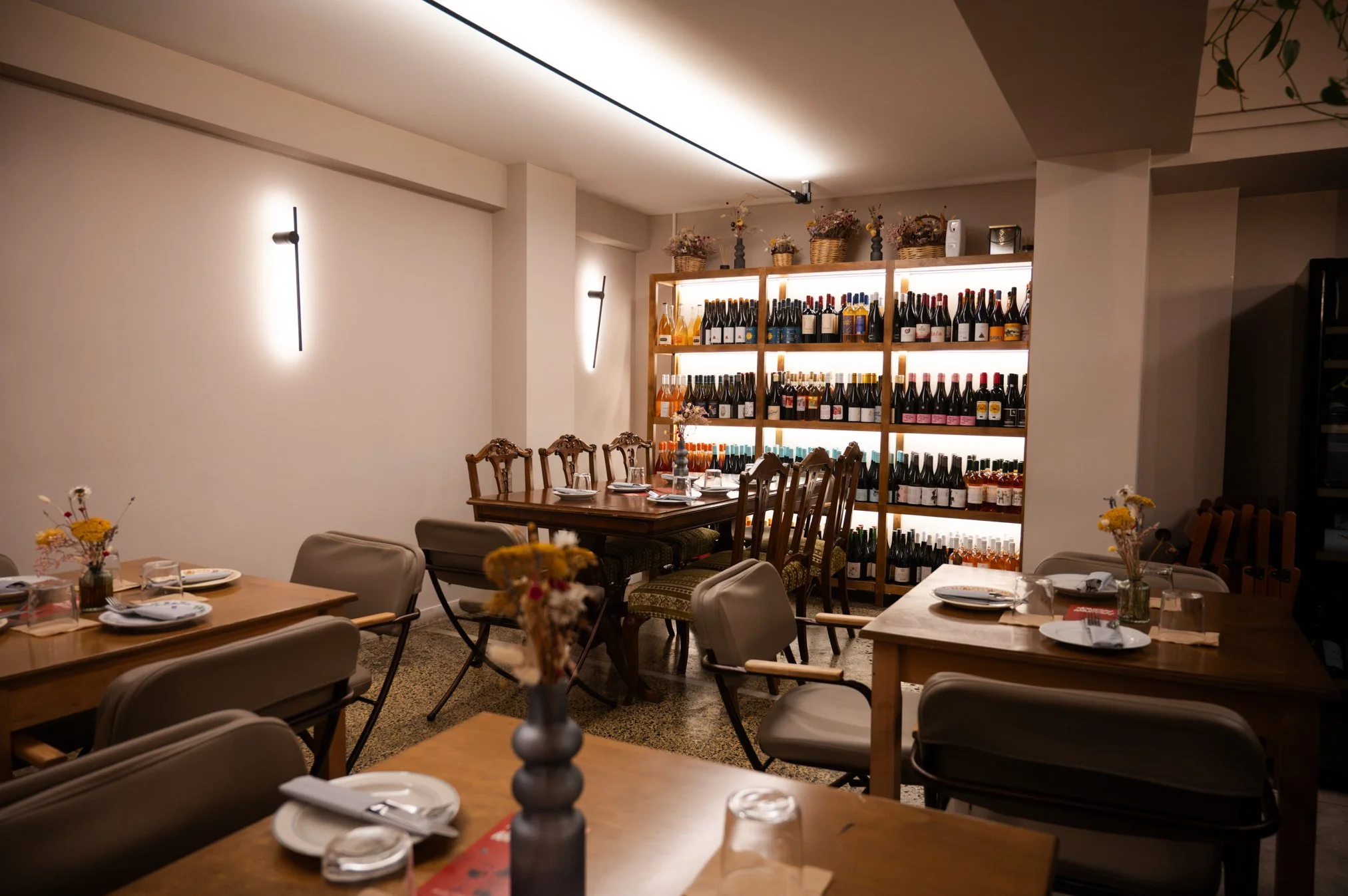 Restaurant with wooden tables, beige chairs, a wine display shelf stocked with various bottles, and small vases of dried flowers on the tables and shelf.