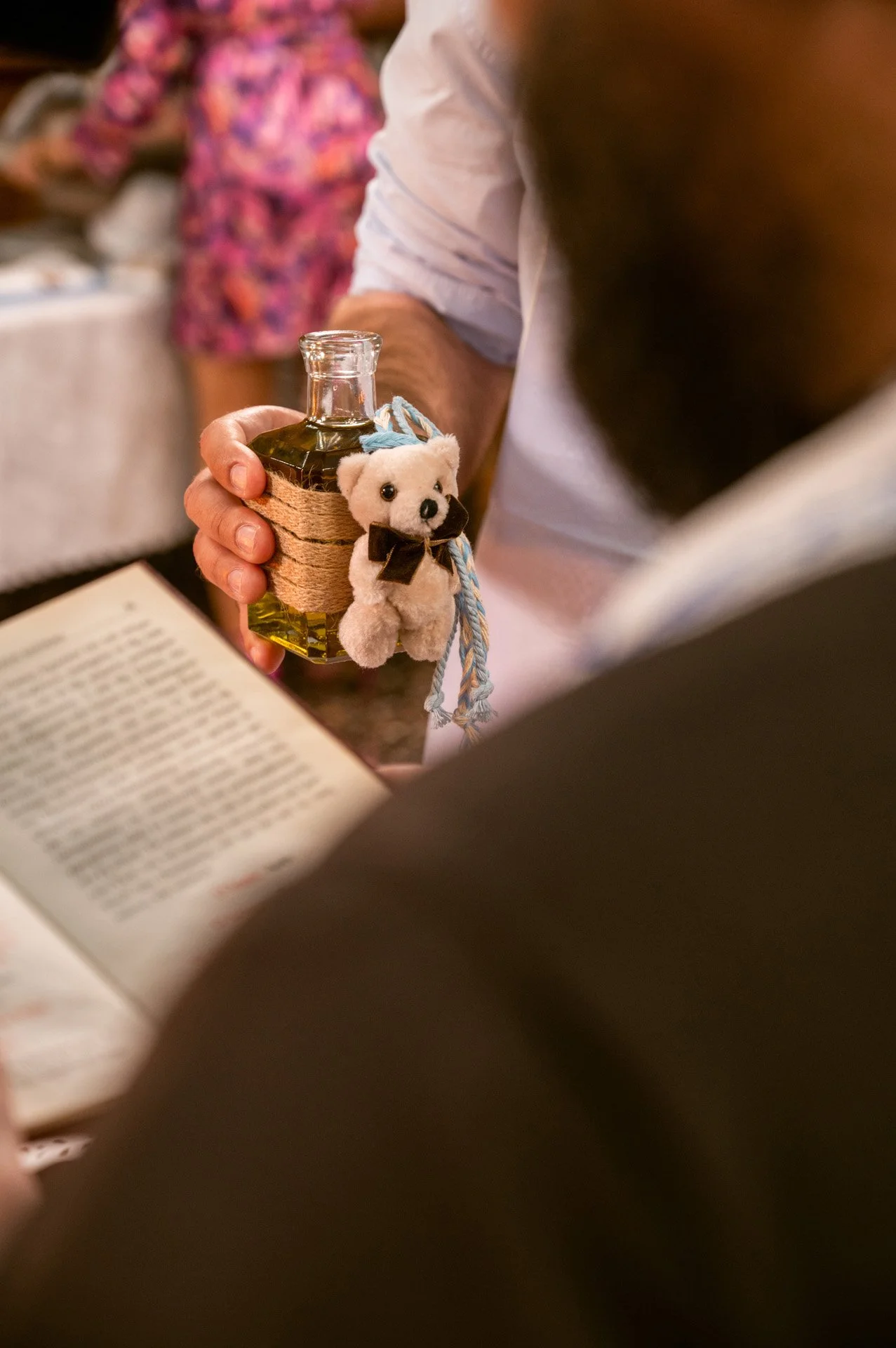 Man holding a small bottle with a teddy bear charm attached, with a person reading a book nearby and a woman in a floral dress in the background.