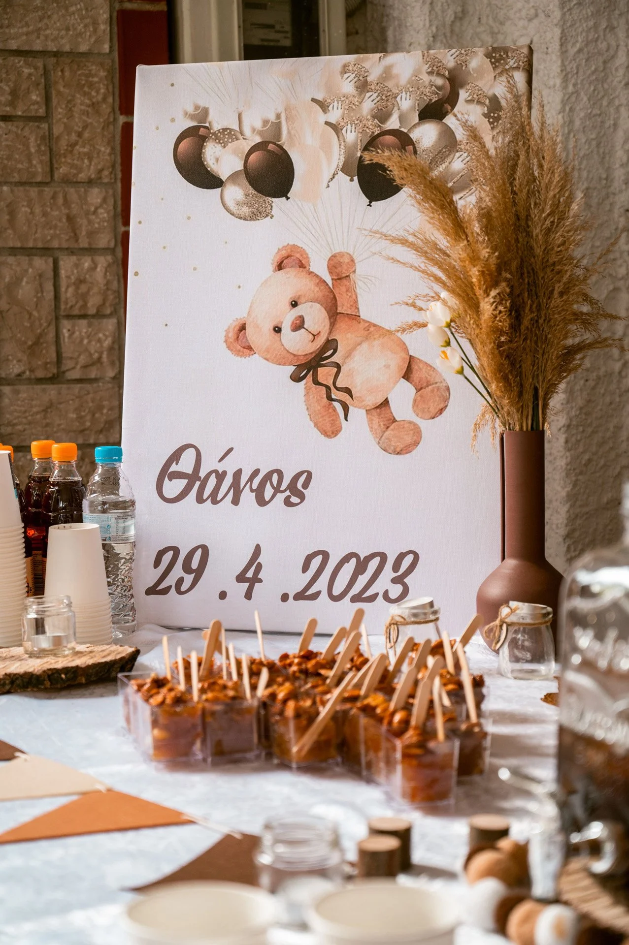 Party table with a sign reading 'Óraros 29.4.2023', featuring a teddy bear illustration, balloons, and decorative dried plants with snacks and beverages.
