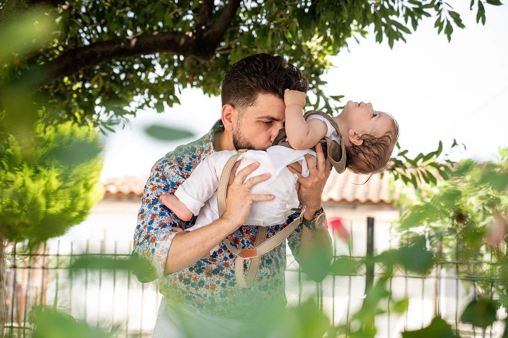 A man holding a child and kissing the child's forehead outdoors under a tree.