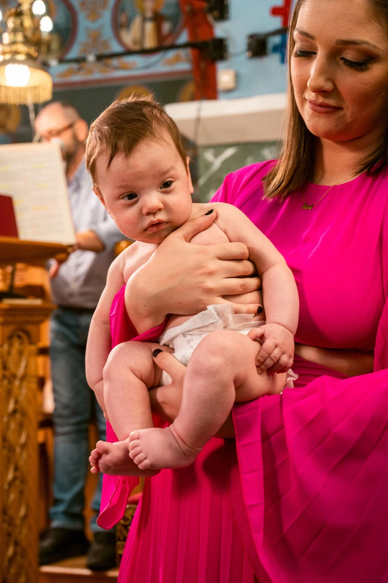 A woman in a bright pink dress holds a baby in a church. The baby looks directly at the camera while the woman looks down. In the background, a man with glasses reads a book.
