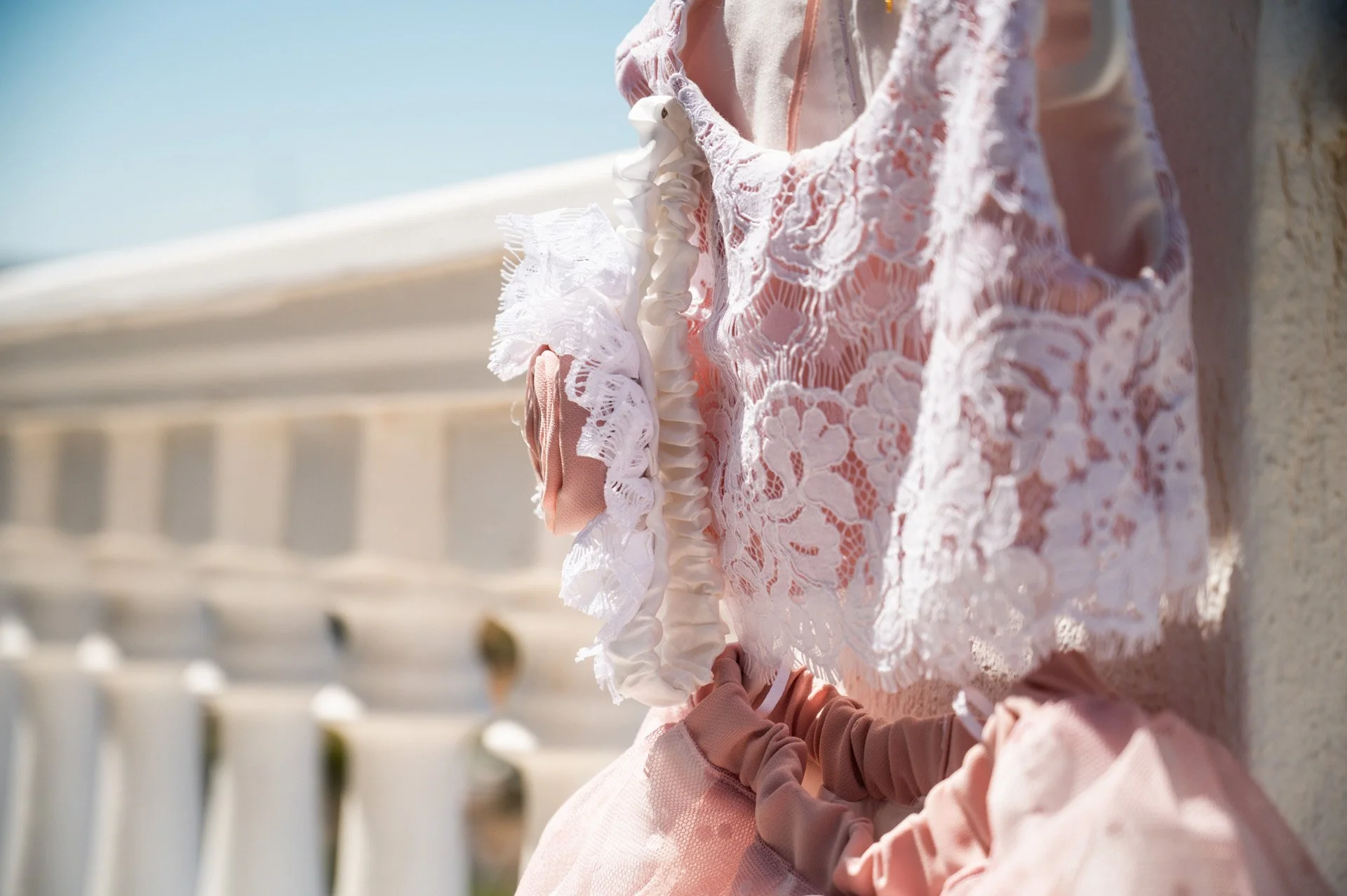 Close-up of a woman wearing pink and white lace clothing, holding a romantic pink and white lace and fabric bouquet, sitting outdoors near a stone railing.