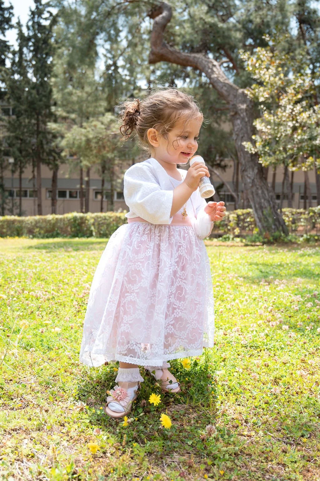 Young girl standing in a grassy area with trees, wearing a white dress with lace and a white cardigan, holding an ice cream cone, smiling.