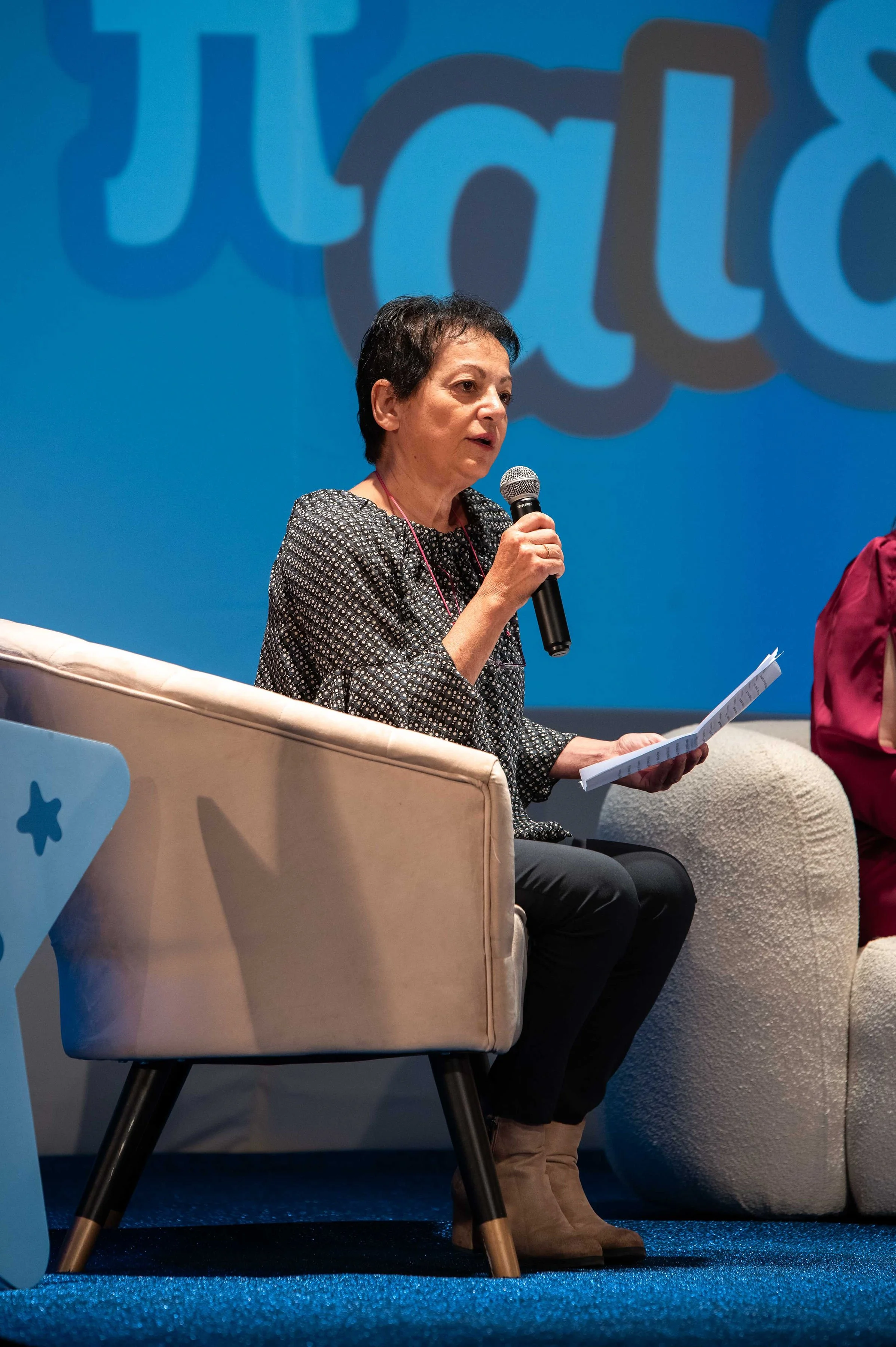 A woman with short dark hair sitting on an armchair, holding a microphone in her right hand and a sheet of paper in her left, speaking at an event with a blue backdrop and partially visible large text.