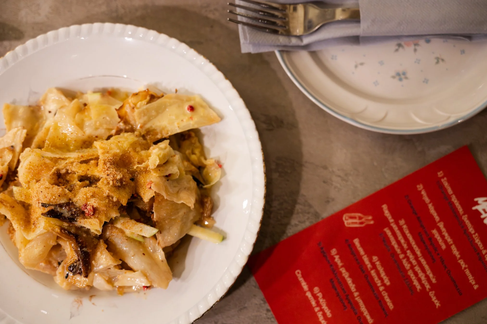 A plate of chicken and mushroom lasagna on a white paper dinner plate. A fork wrapped with a napkin sits on a nearby plate, and a red menu or advertisement is partially visible on a brown table.