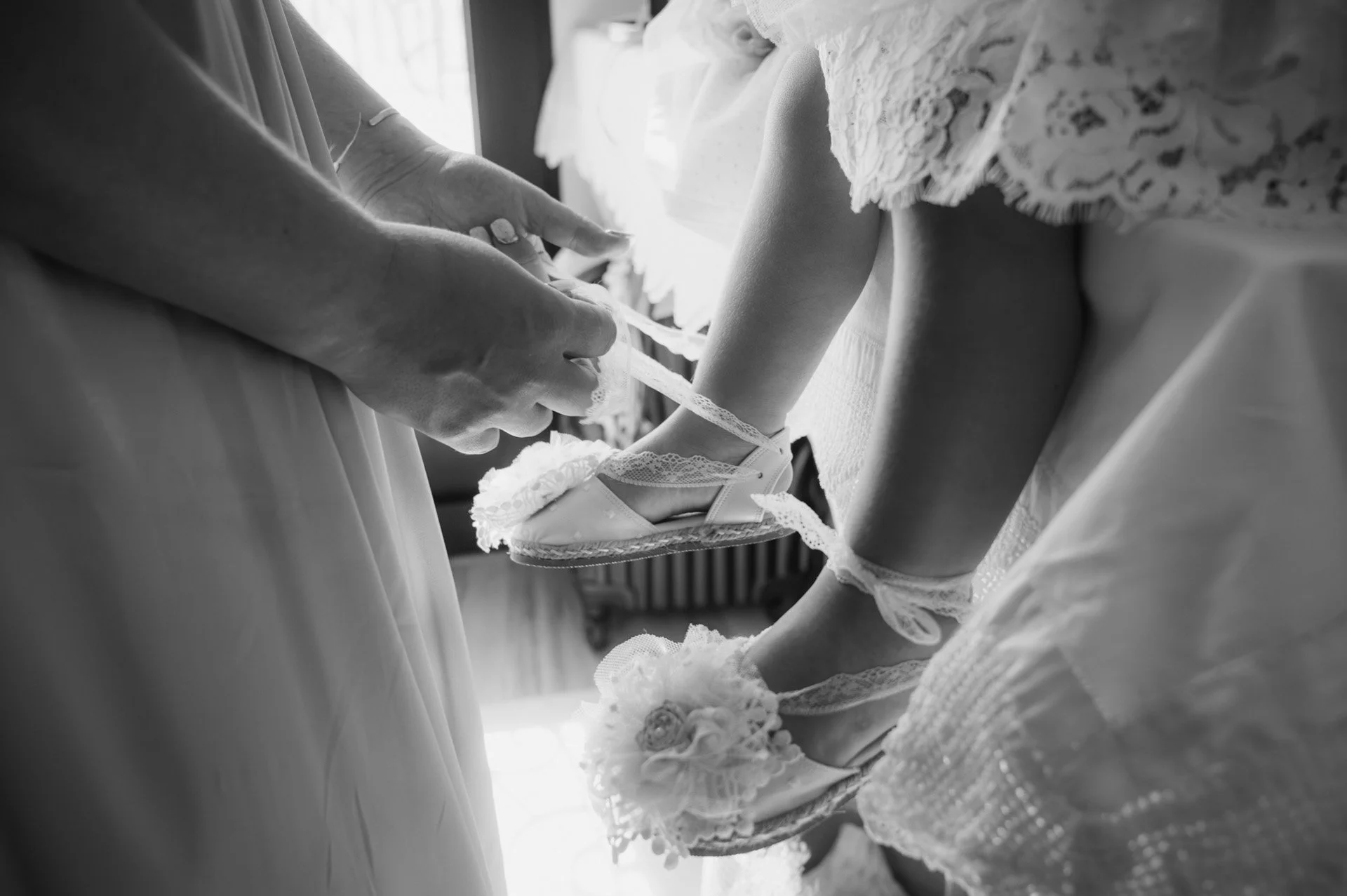 A woman adjusts the shoes on a young girl, both dressed in white with lace details, likely preparing for a special event or wedding.