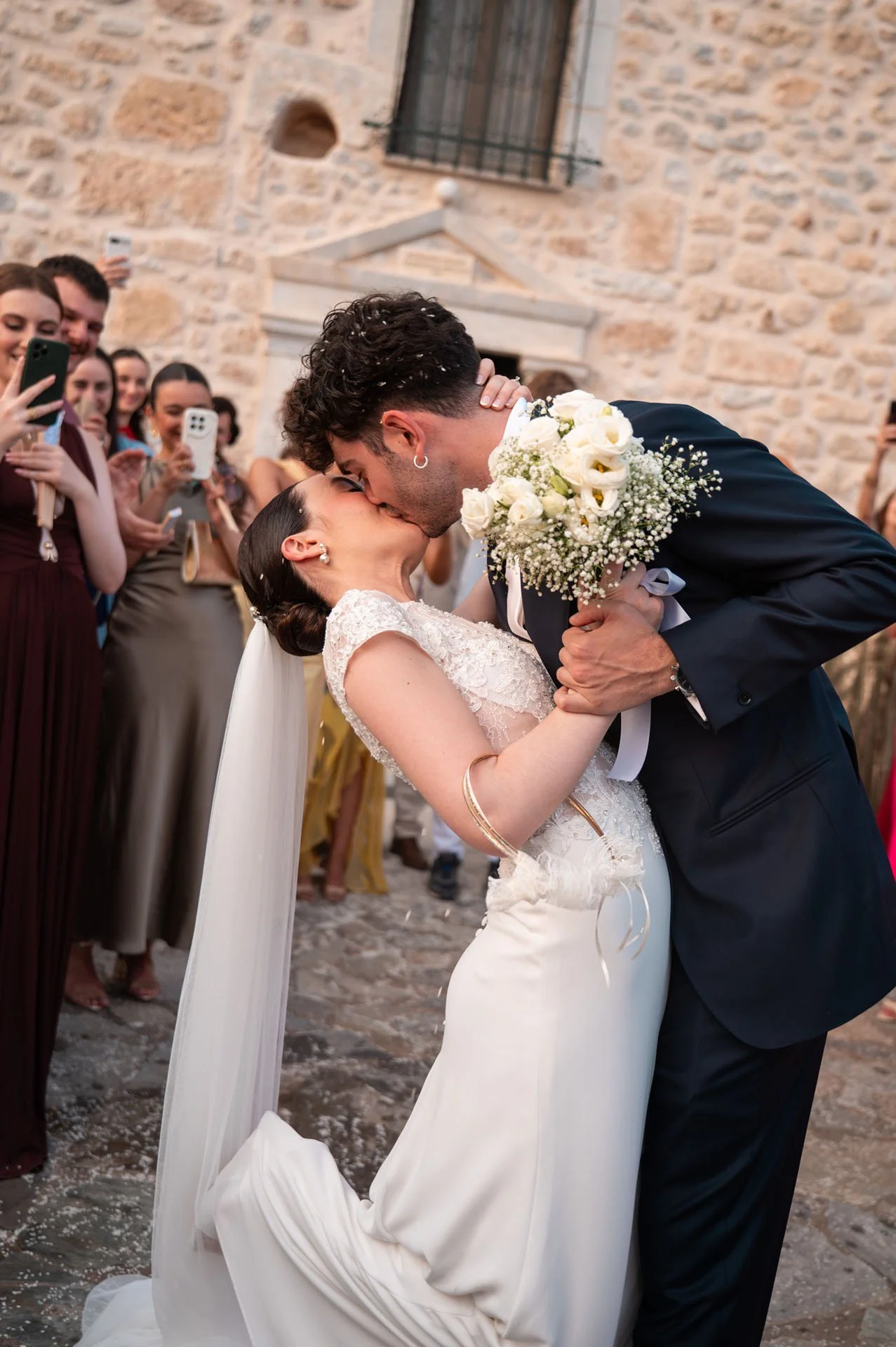 A couple kissing during their wedding ceremony outside. The bride is in a white gown holding a bouquet, and the groom is in a dark suit. Guests are taking photos and cheering in the background, with a stone building behind them.