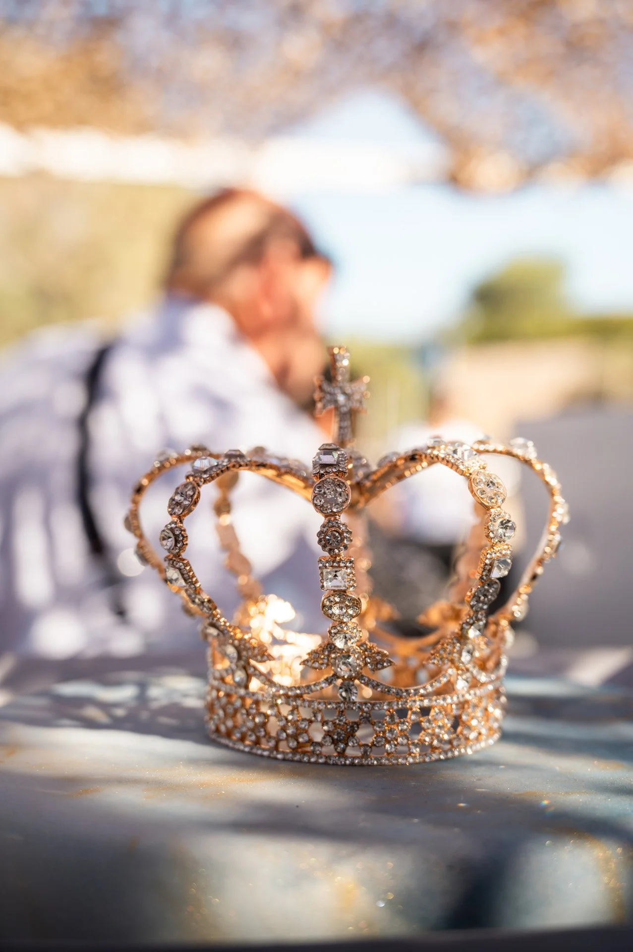 Close-up of a jeweled crown with a cross on top, placed on a reflective surface outdoors, with a person blurred in the background.