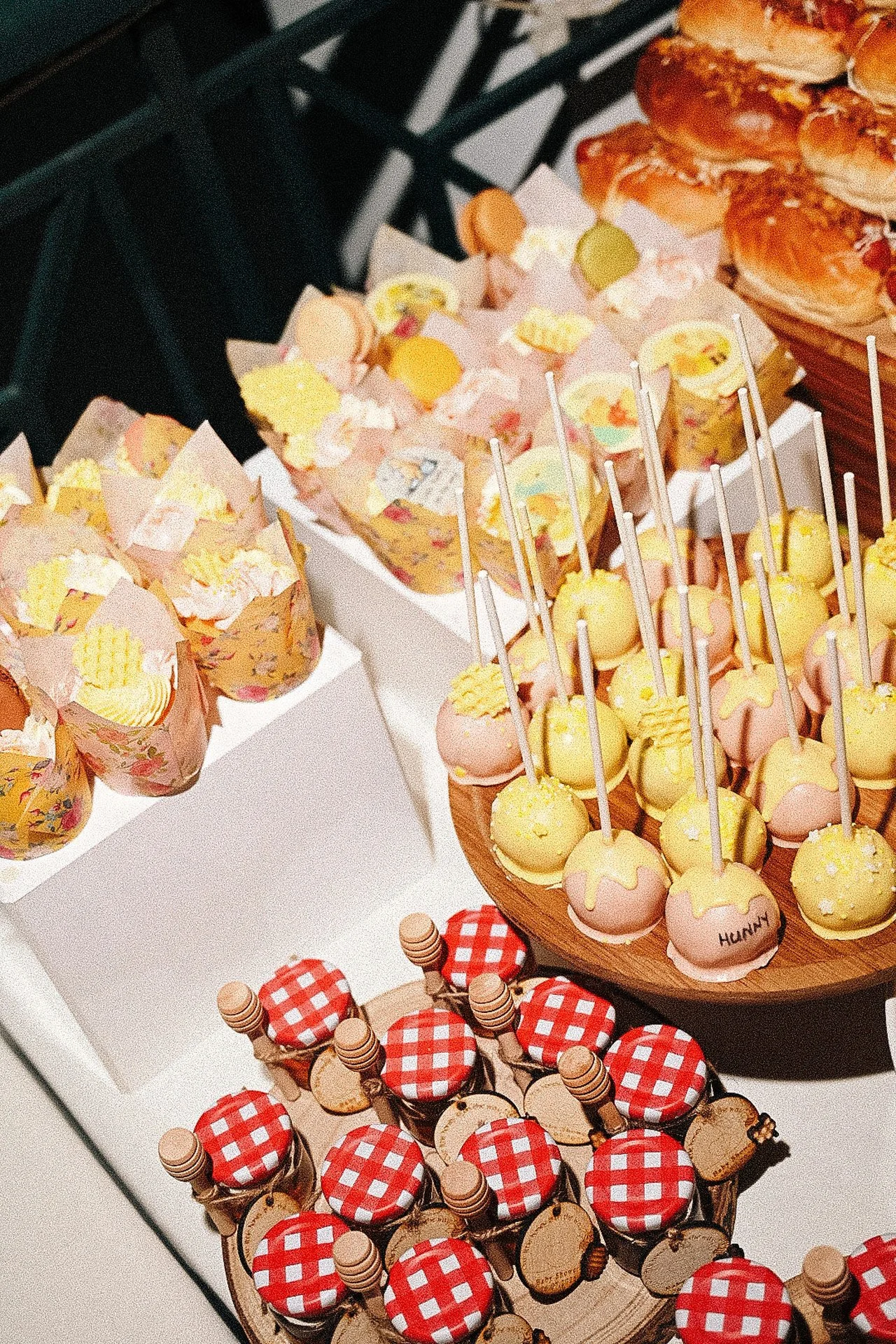 Assorted desserts including cake pops with yellow and pink icing with sticks, small cakes in floral paper wrappers, and jars with red checkered lids on a white table.