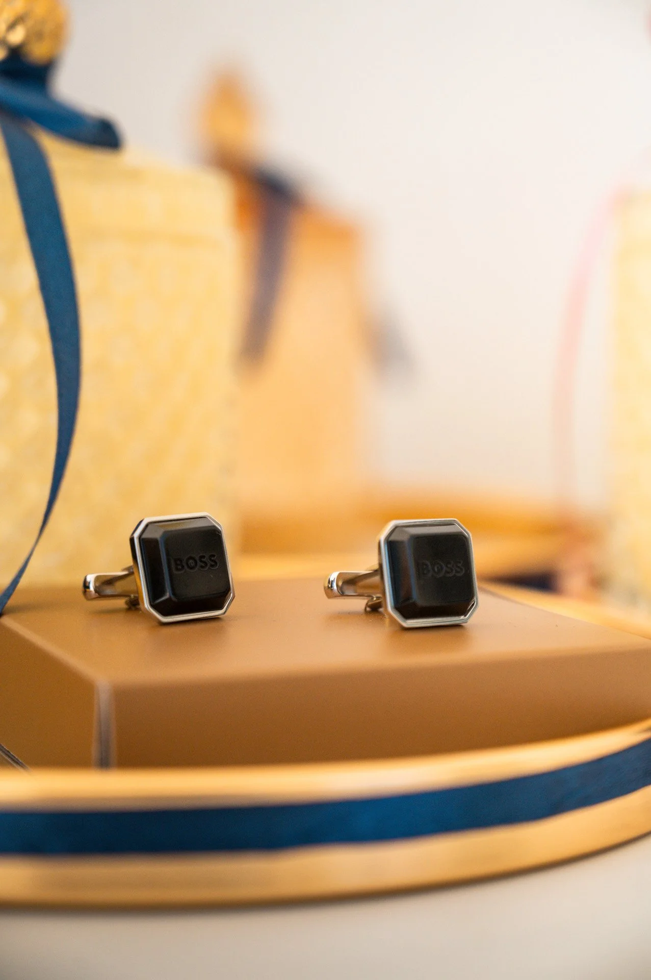 A pair of black and silver Boss cufflinks resting on a golden tray with navy blue trim, blurred background.